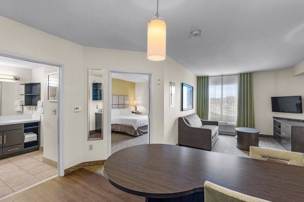 Habitación de hotel con una mesa redonda para comer, un sofá, un televisor y un amplio ventanal con cortinas.