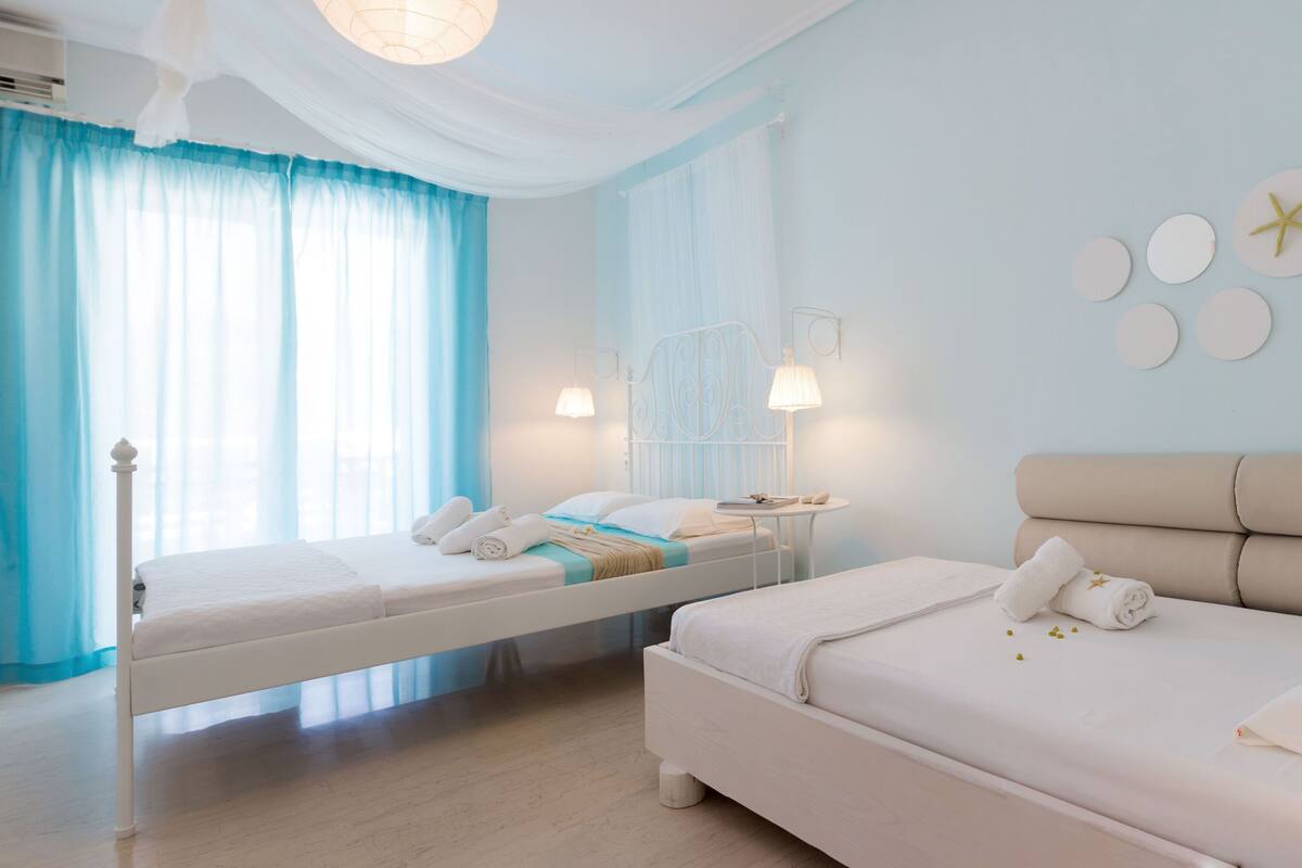 A hotel room with two beds, a large window with blue curtains, a wall-mounted lamp, and a decorative starfish on the wall.