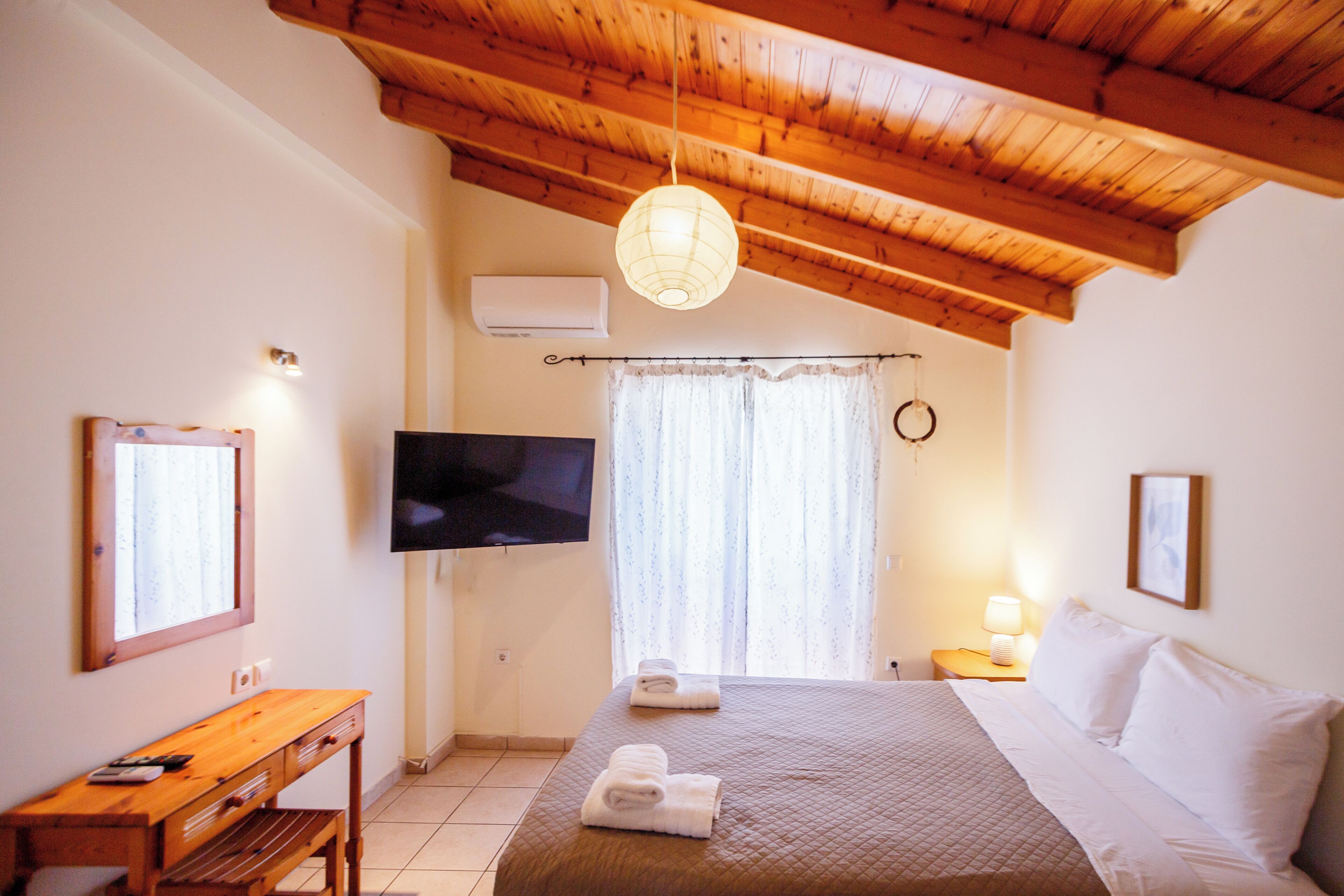 Ein Schlafzimmer mit Holzboden, einem Bett mit weißen Leinentüchern, einem Schreibtisch mit Stuhl, einem an der Wand montierten Flachbildfernseher und einem Fenster mit Vorhängen.