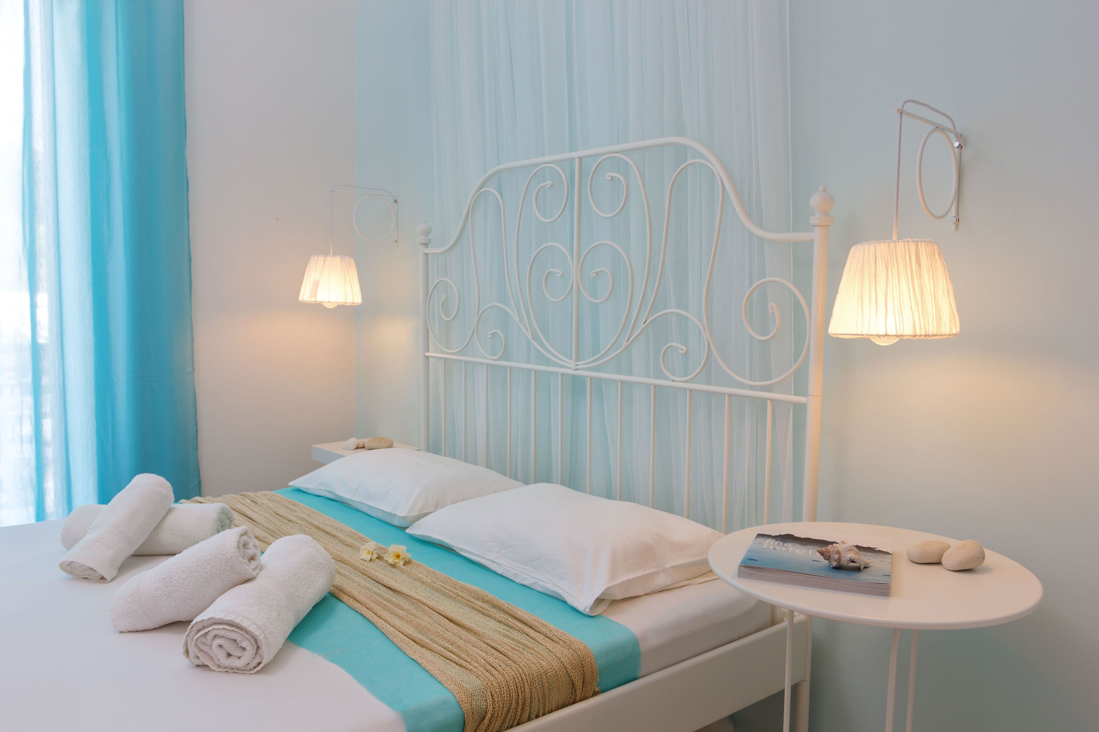 A neatly made bed with rolled towels, a bedside table with a book and two stones, and wall-mounted lamps.