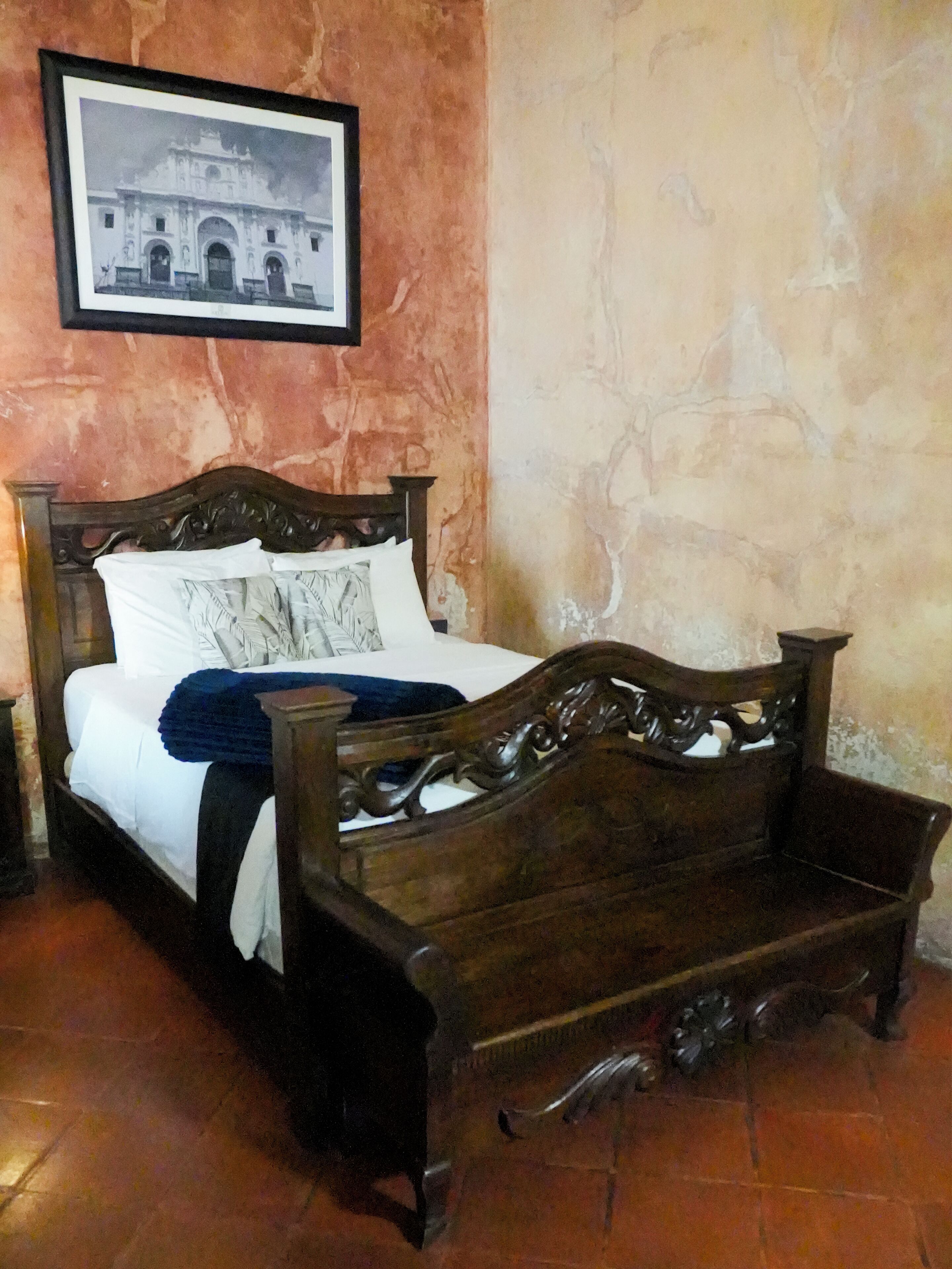 Uma cama com cabeceira de madeira, um quadro na parede e chão de azulejos.