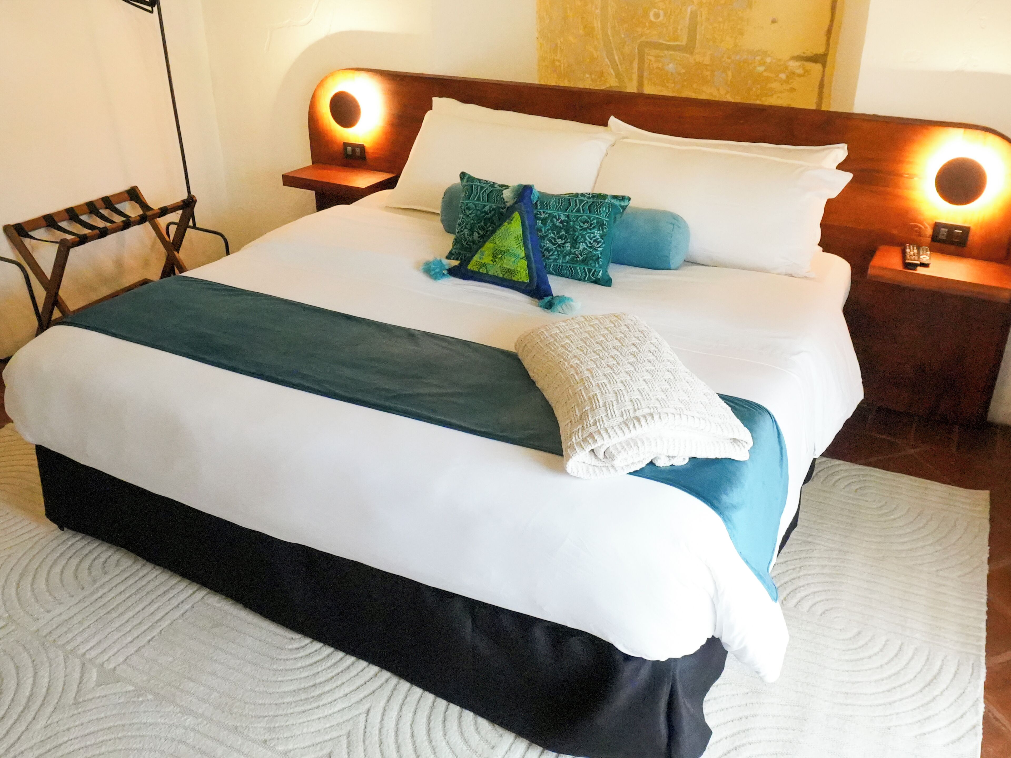Ein ordentlich bezogenes Bett mit weißer und türkiser Decke, zwei Kissen und einer gefalteten Decke. Zwei Nachttische mit beleuchteten Lampen sind vorhanden.