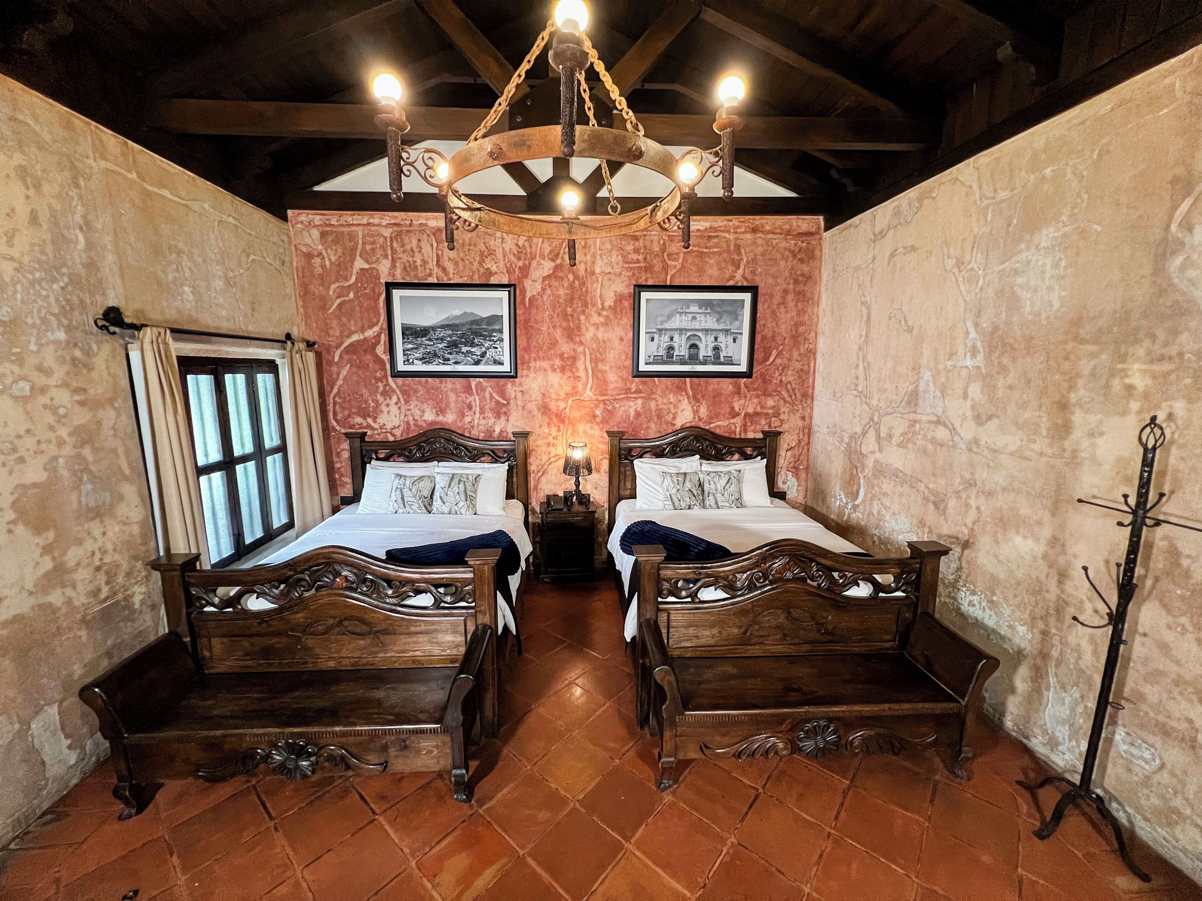 A room with two wooden beds, a chandelier, a window with curtains, and two framed pictures on the wall.
