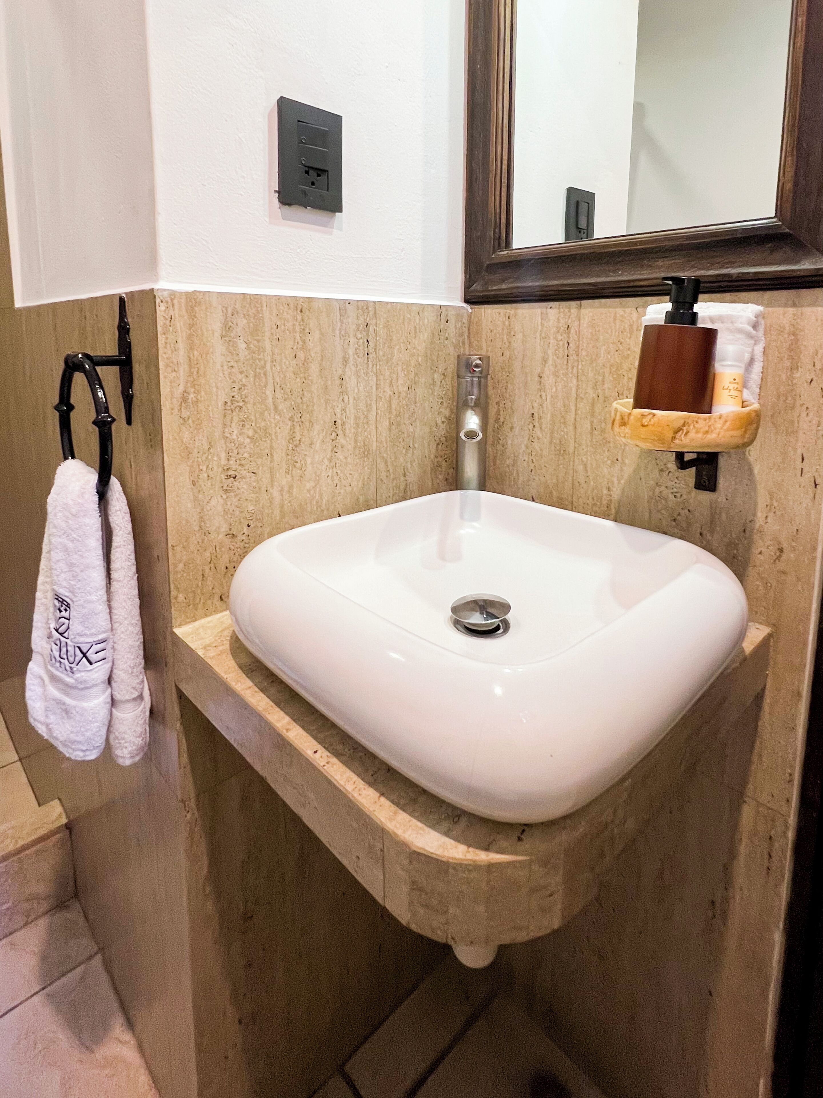 A bathroom with a white sink, a towel rack with a white towel, a wooden shelf with toiletries, and a mirror above the sink.