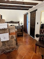 A hotel room with a stone wall, a bed with a wooden headboard, a wooden chair, a desk with a lamp, and a door with a mirror.