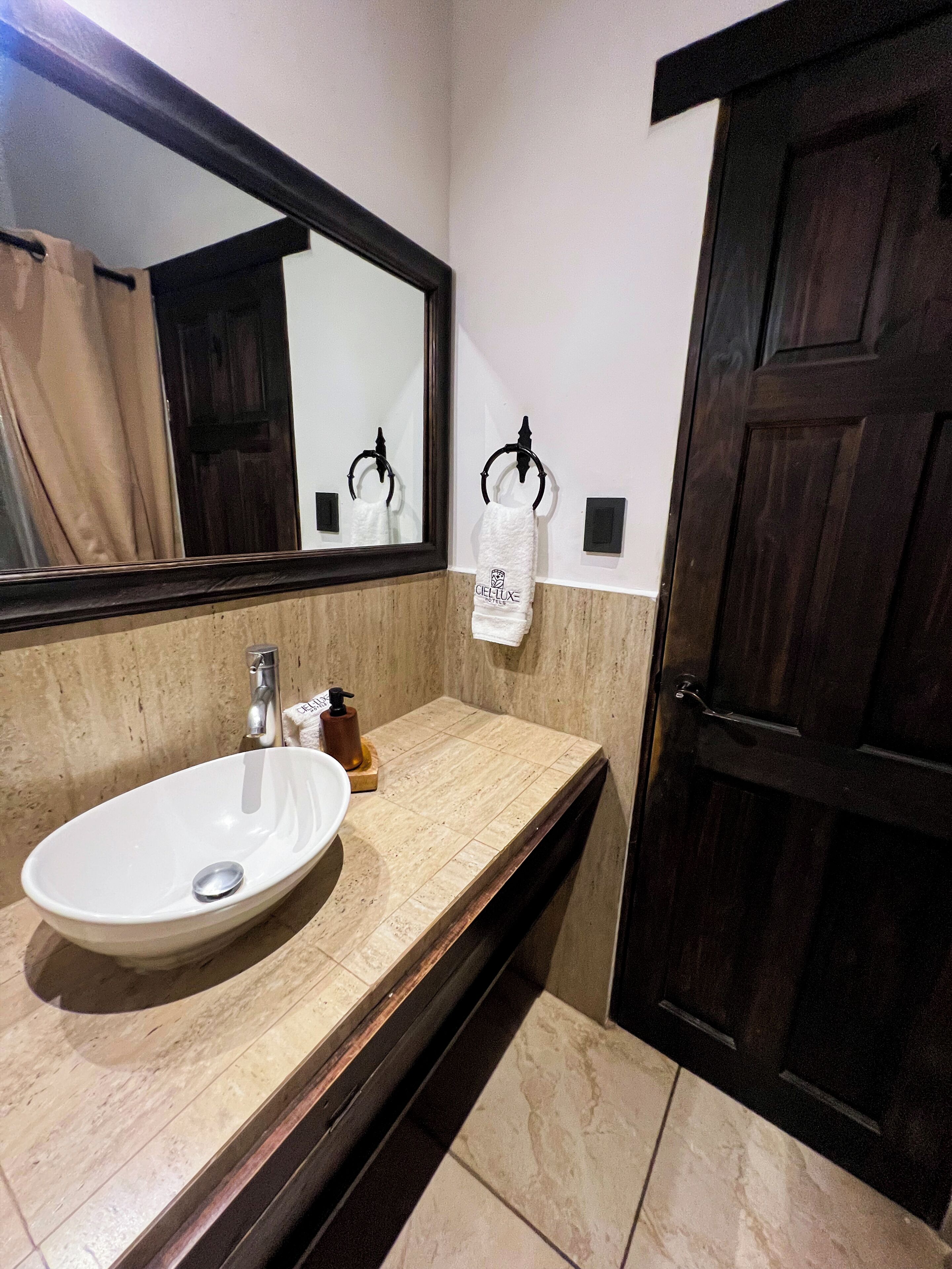 Un baño con lavamanos blanco, un espejo, toallero y una puerta de madera oscura.