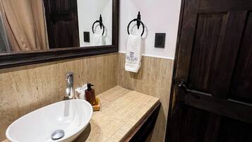Un baño con lavamanos blanco, un espejo, toallero y una puerta de madera oscura.