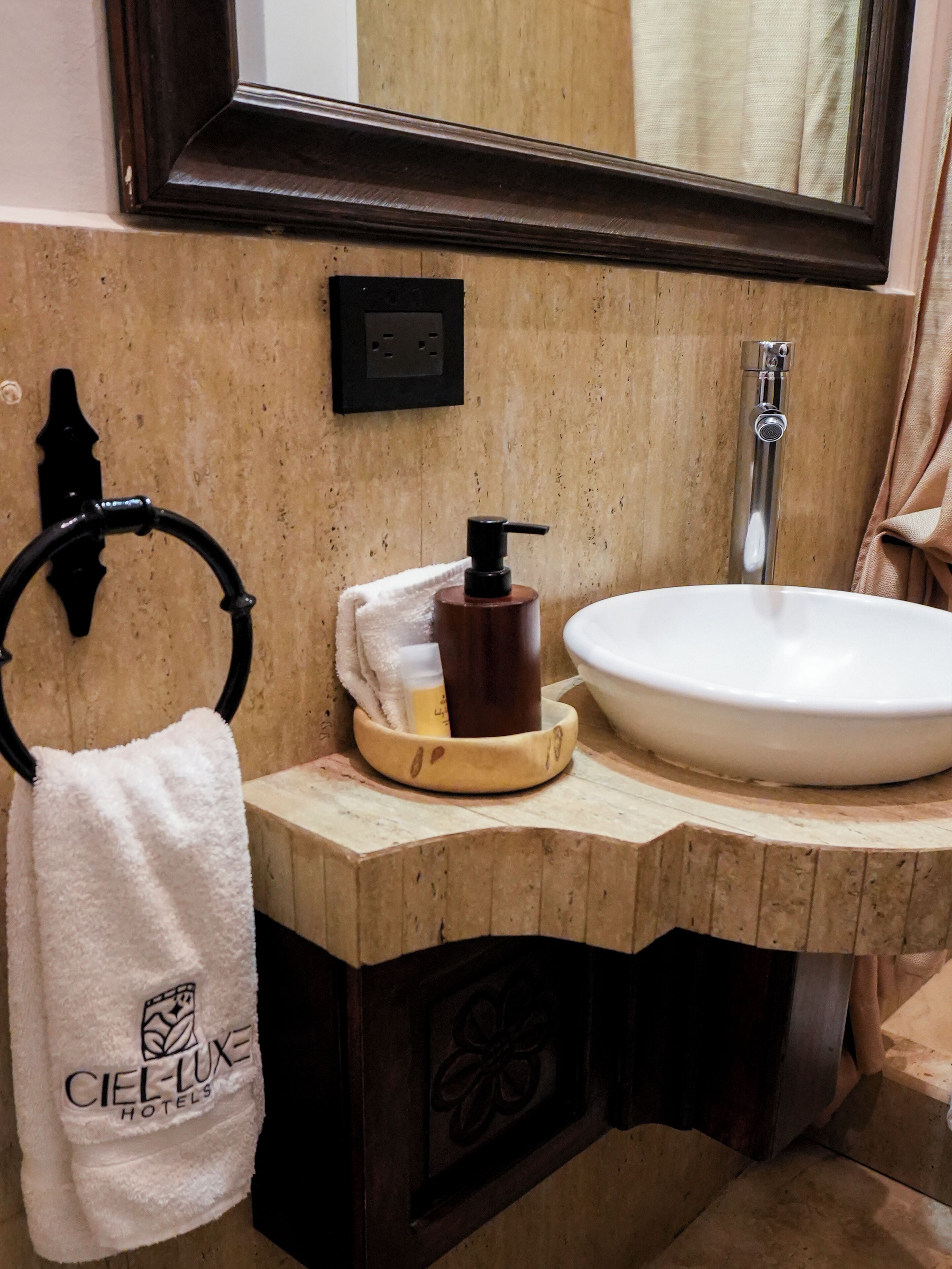 A bathroom with a wooden vanity, a white sink, a mirror, a towel rack, and a soap dispenser.