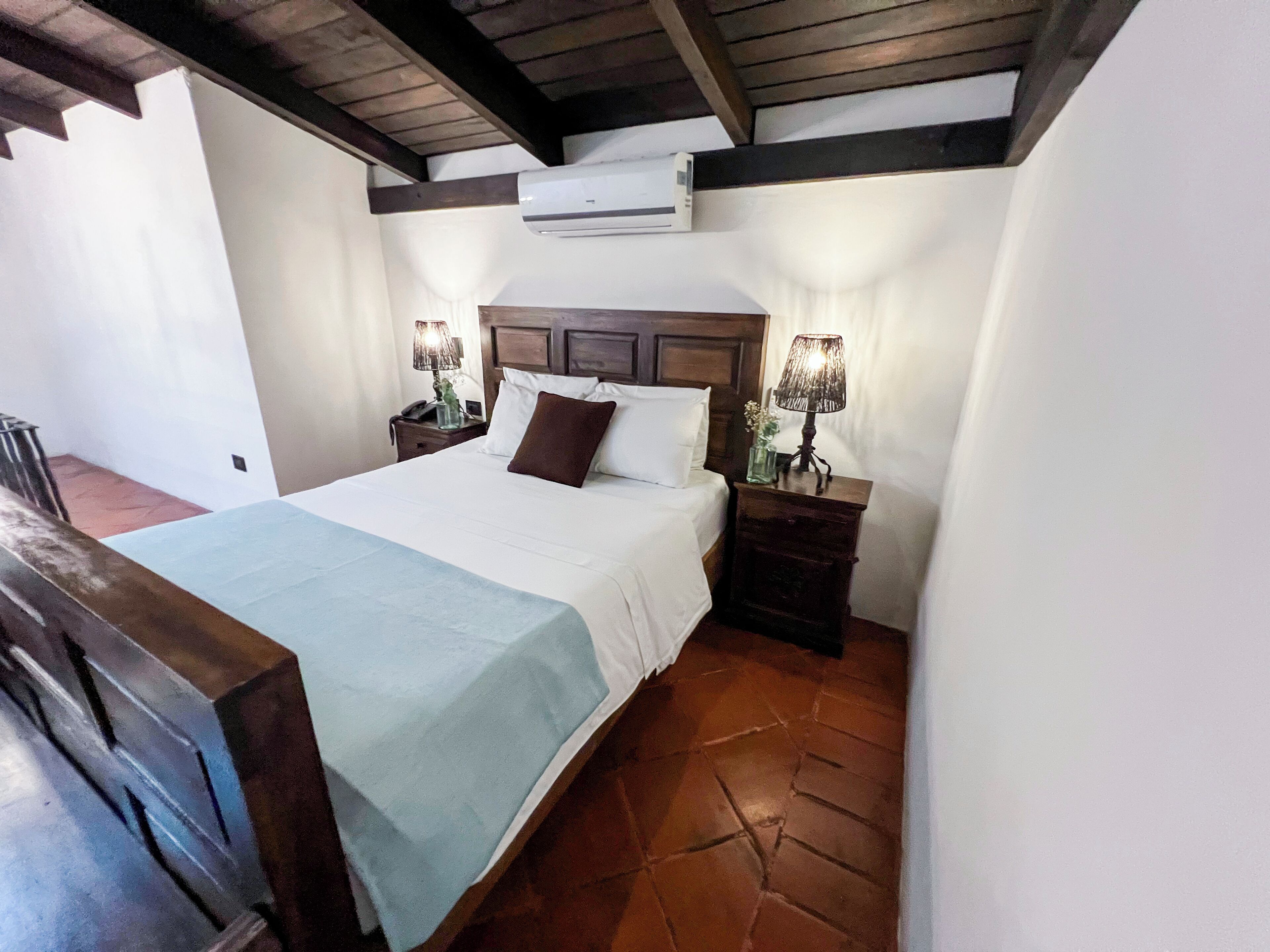 A hotel room with a bed, nightstands, lamps, an air conditioning unit, and a tiled floor.
