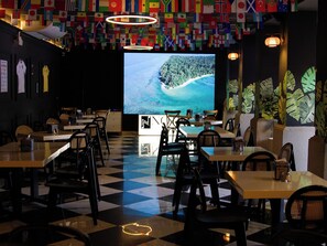 Un restaurante con piso a cuadros, mesas, sillas, una gran pantalla que muestra una playa tropical y una pared decorada con varias banderas nacionales.