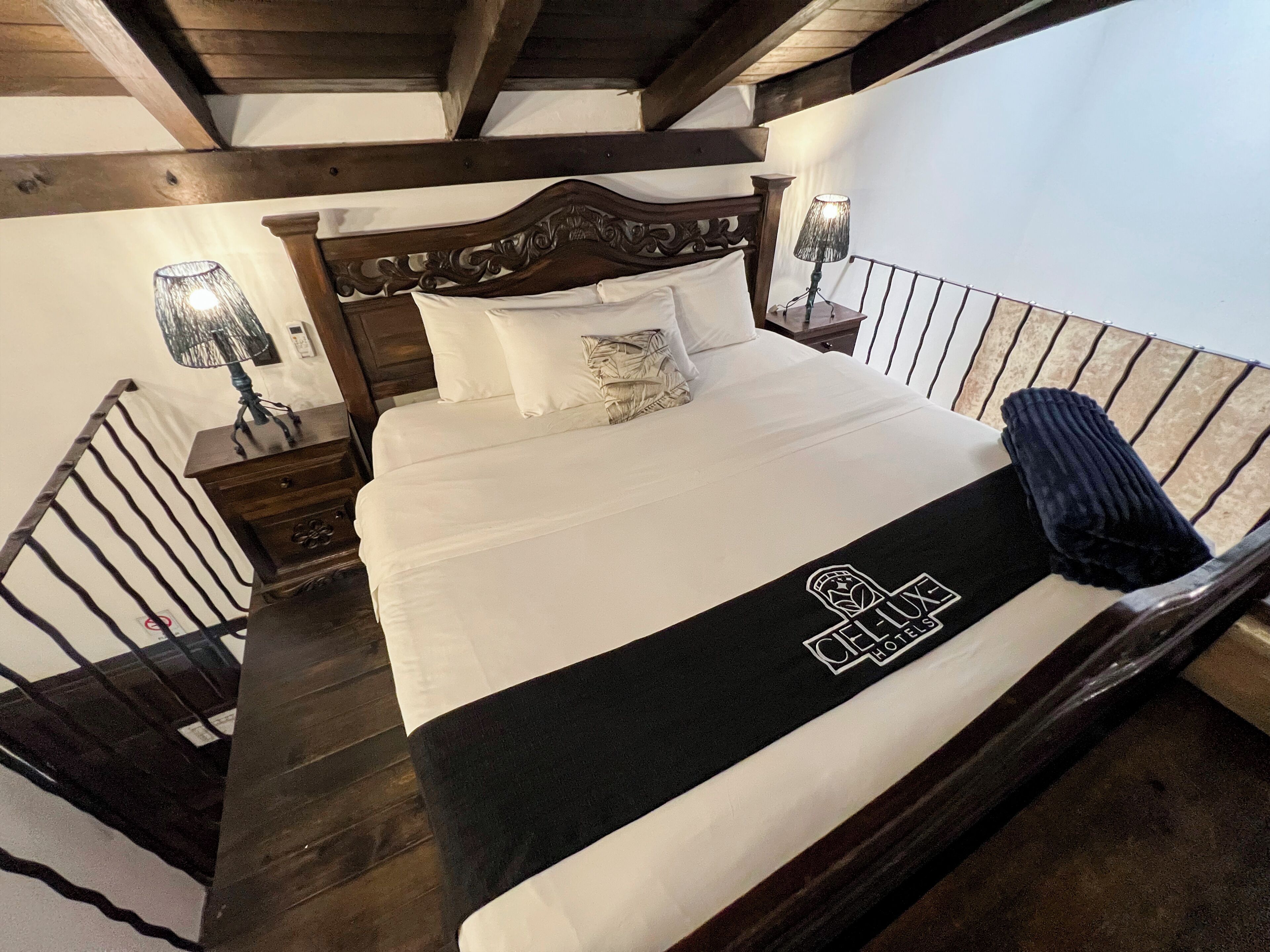 Habitación de hotel con cabecera de madera, cama con ropa de cama blanca, manta negra con un logotipo, mesita de noche con lámpara y una escalera.