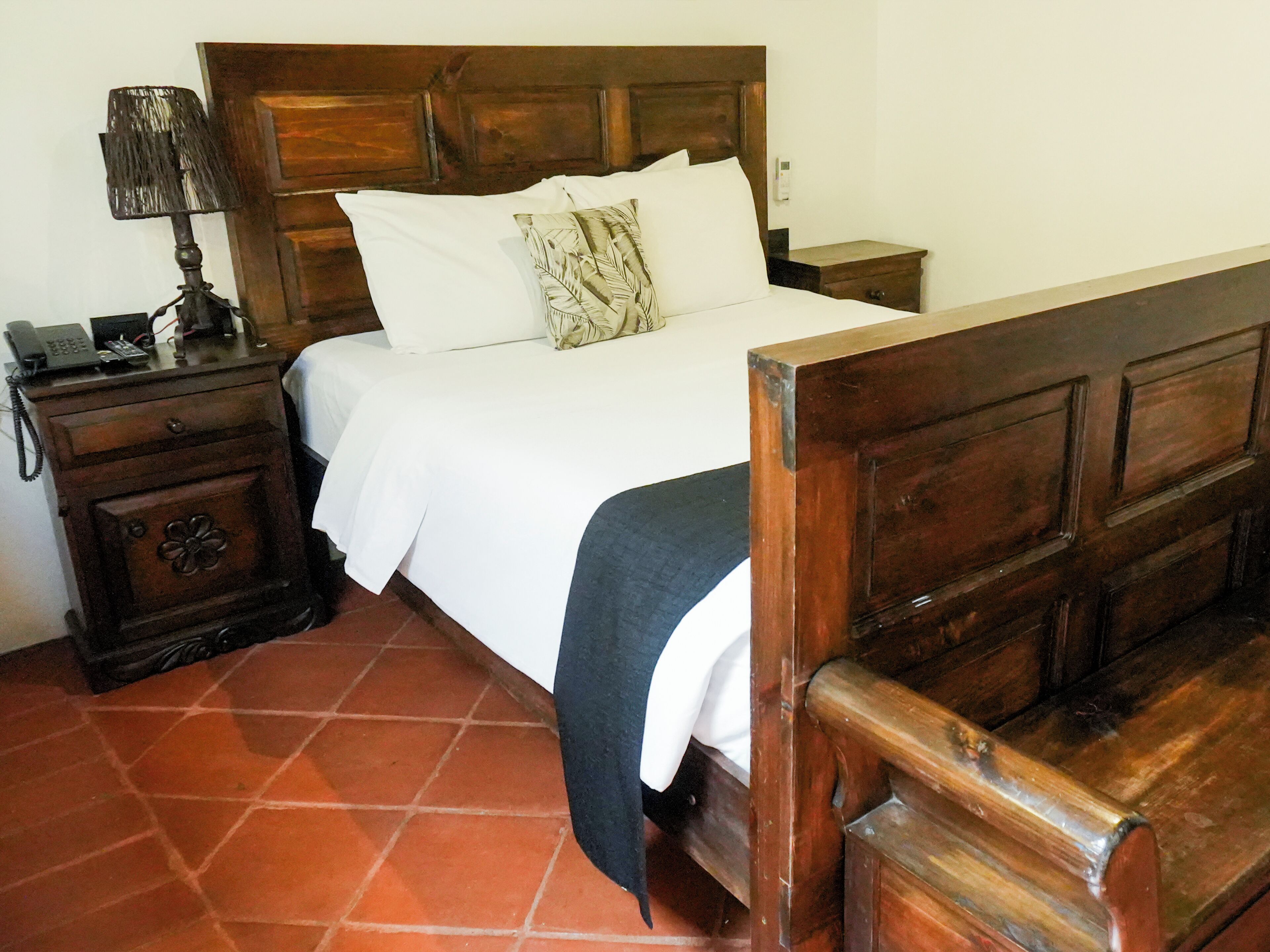 Una cama de madera con ropa blanca, una mesita de noche con lámpara y un mueble con cajones.