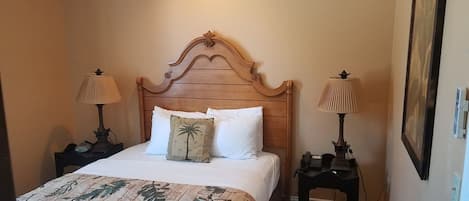 Une chambre d’hôtel avec une tête de lit en bois, deux lampes de chevet, un lit recouvert d’une couvre-lit à motifs floraux et une table de chevet avec une lampe.