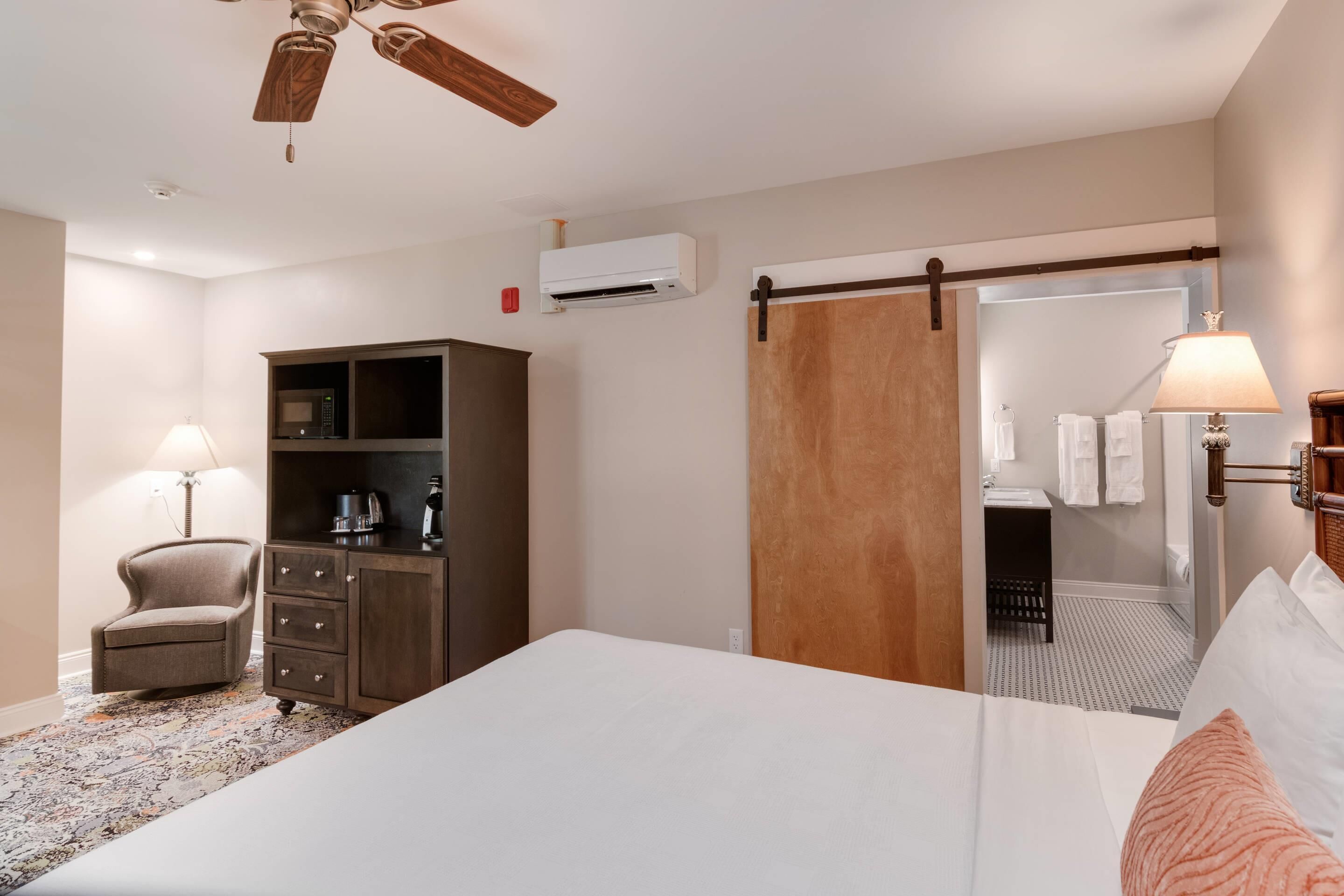 A hotel room with a bed, a desk with a lamp, a chair, a wardrobe, an air conditioning unit, and a sliding door to a bathroom.