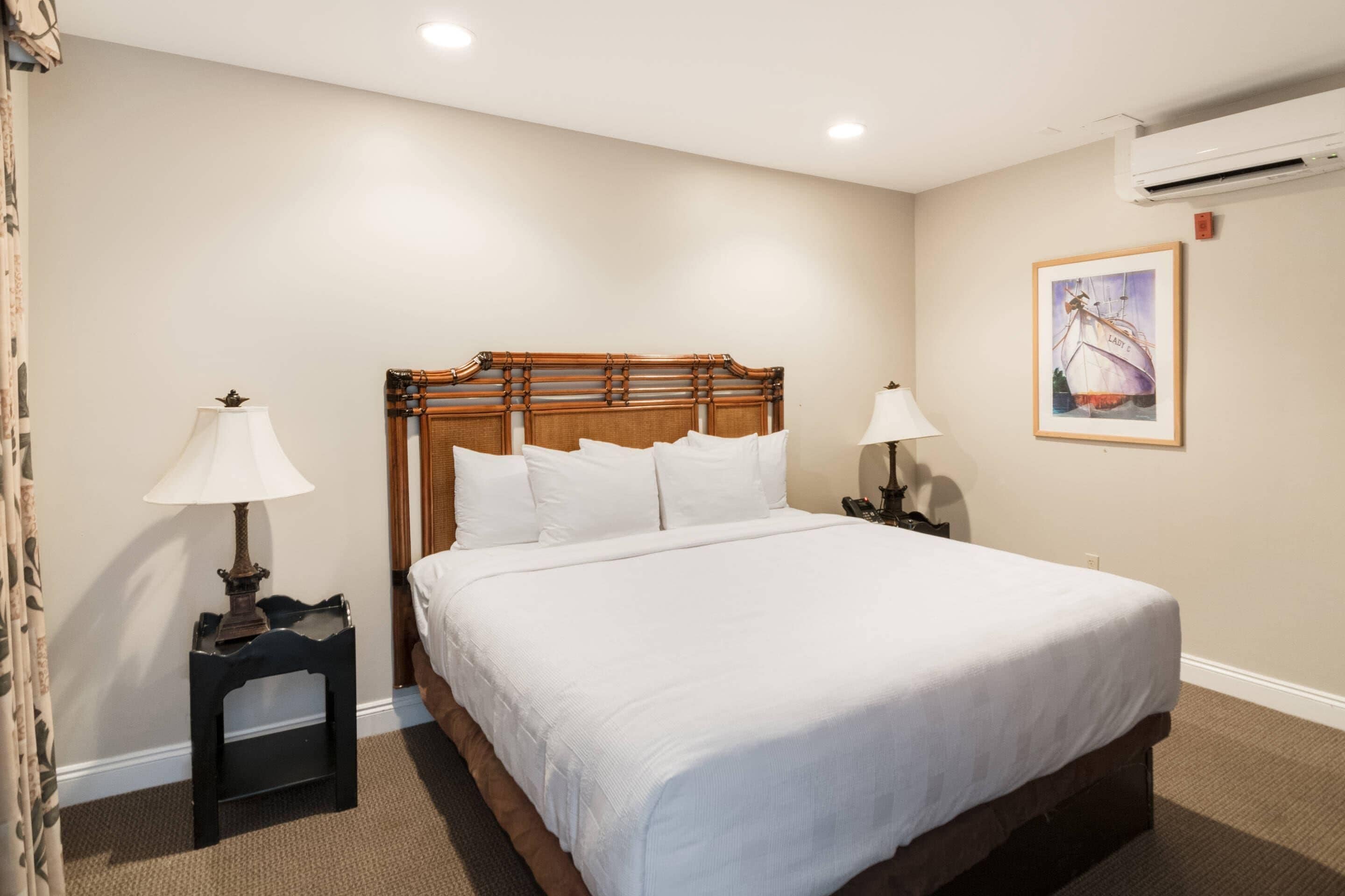 A neatly made bed with a wooden headboard, two bedside tables with lamps, a framed picture on the wall, and an air conditioning unit.
