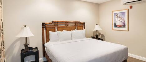 A neatly made bed with a wooden headboard, two bedside tables with lamps, a framed picture on the wall, and an air conditioning unit.