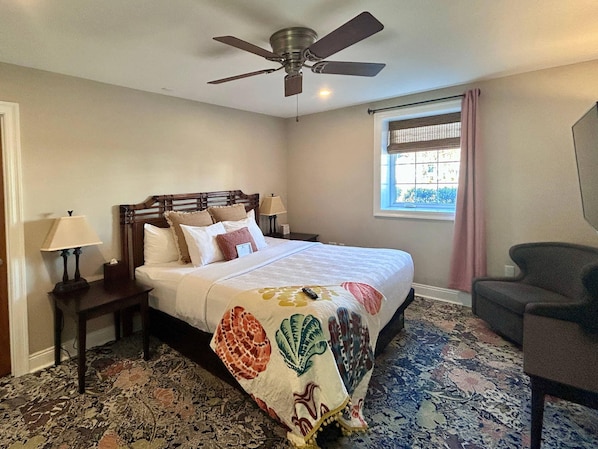Une chambre à coucher comprenant un lit, un ventilateur de plafond, une fenêtre avec des rideaux, une table de chevet avec une lampe et une chaise.