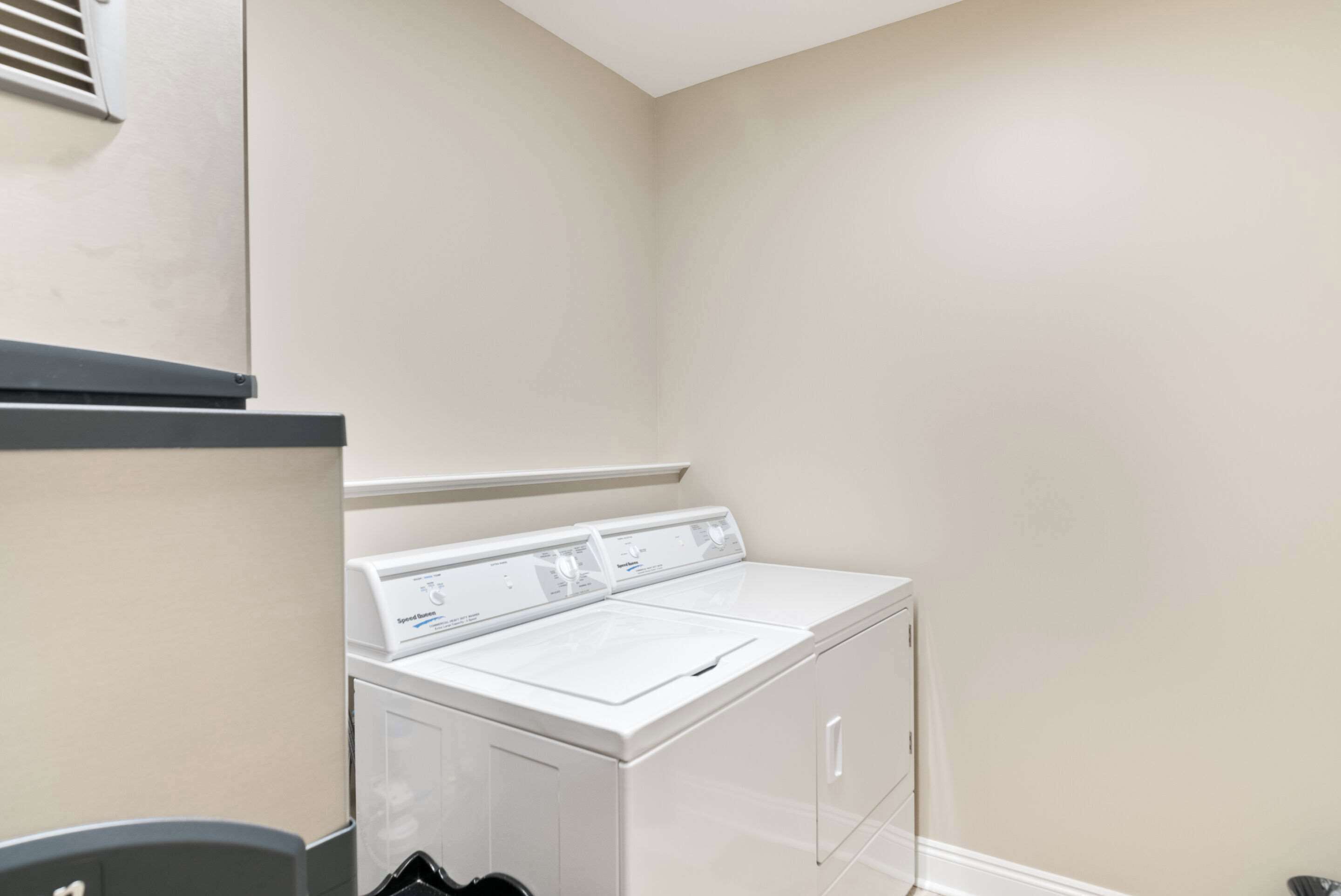 A laundry room with a front-loading washer and dryer, a small trash bin, and a vent on the wall.