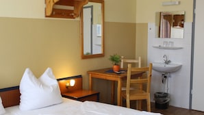 A hotel room with a bed, a bedside table with a lamp, a wooden chair, a small table with a plant, and a bathroom with a sink and mirror. - Pension Schwalbenweg (Schoenefeld)