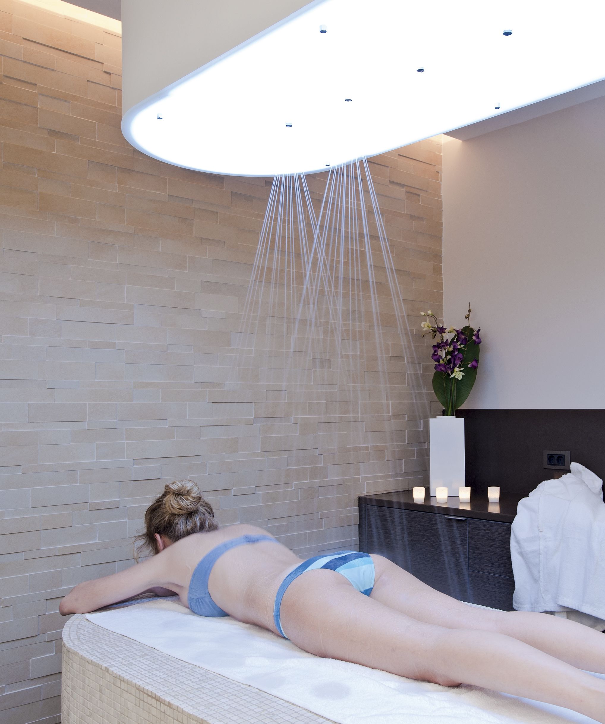 A person lying on a massage table in a spa room with a modern design, including a brick wall and a ceiling light fixture.