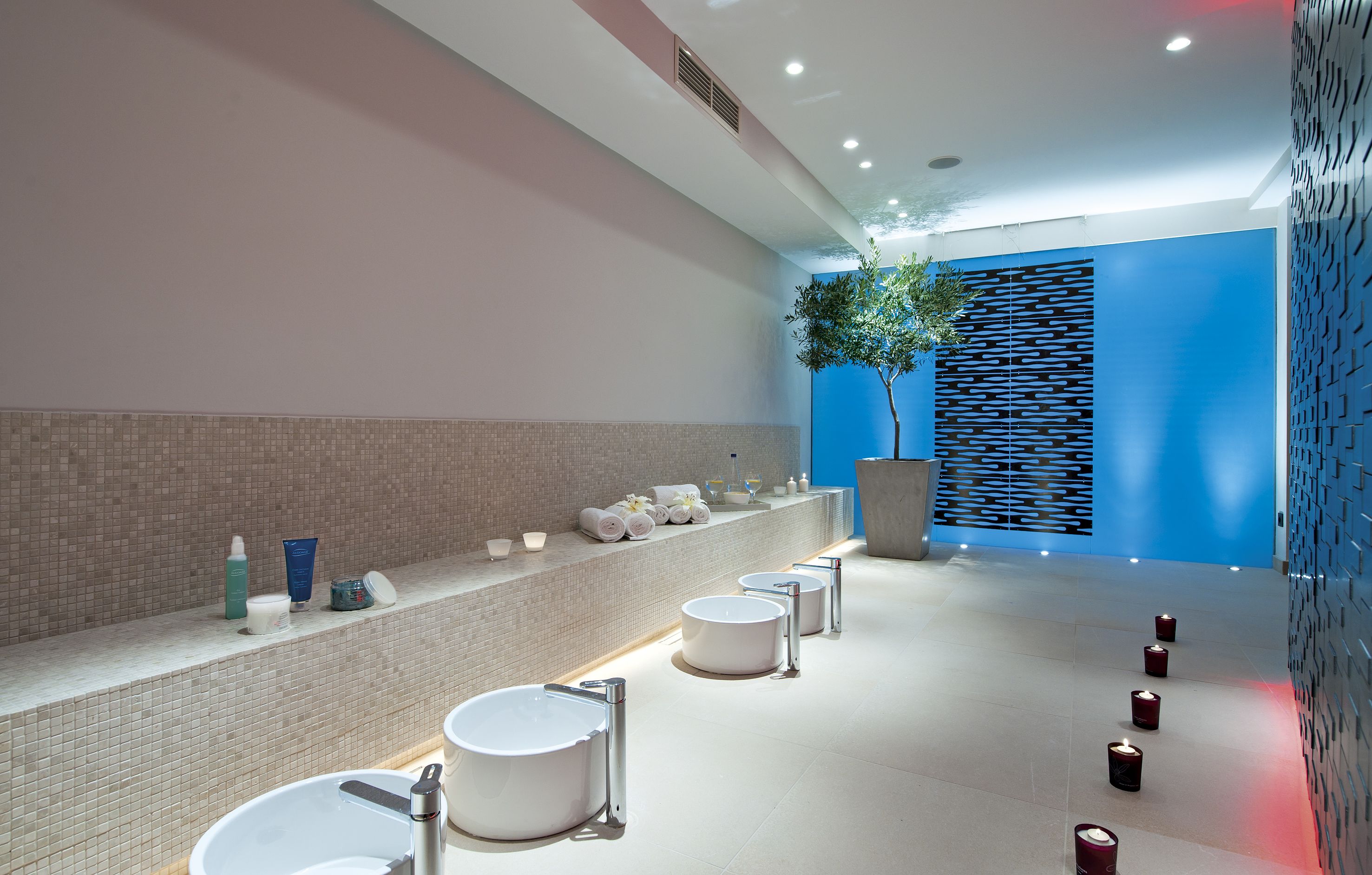 A modern bathroom with multiple sinks, a long vanity with toiletries, and a decorative wall with a tree.