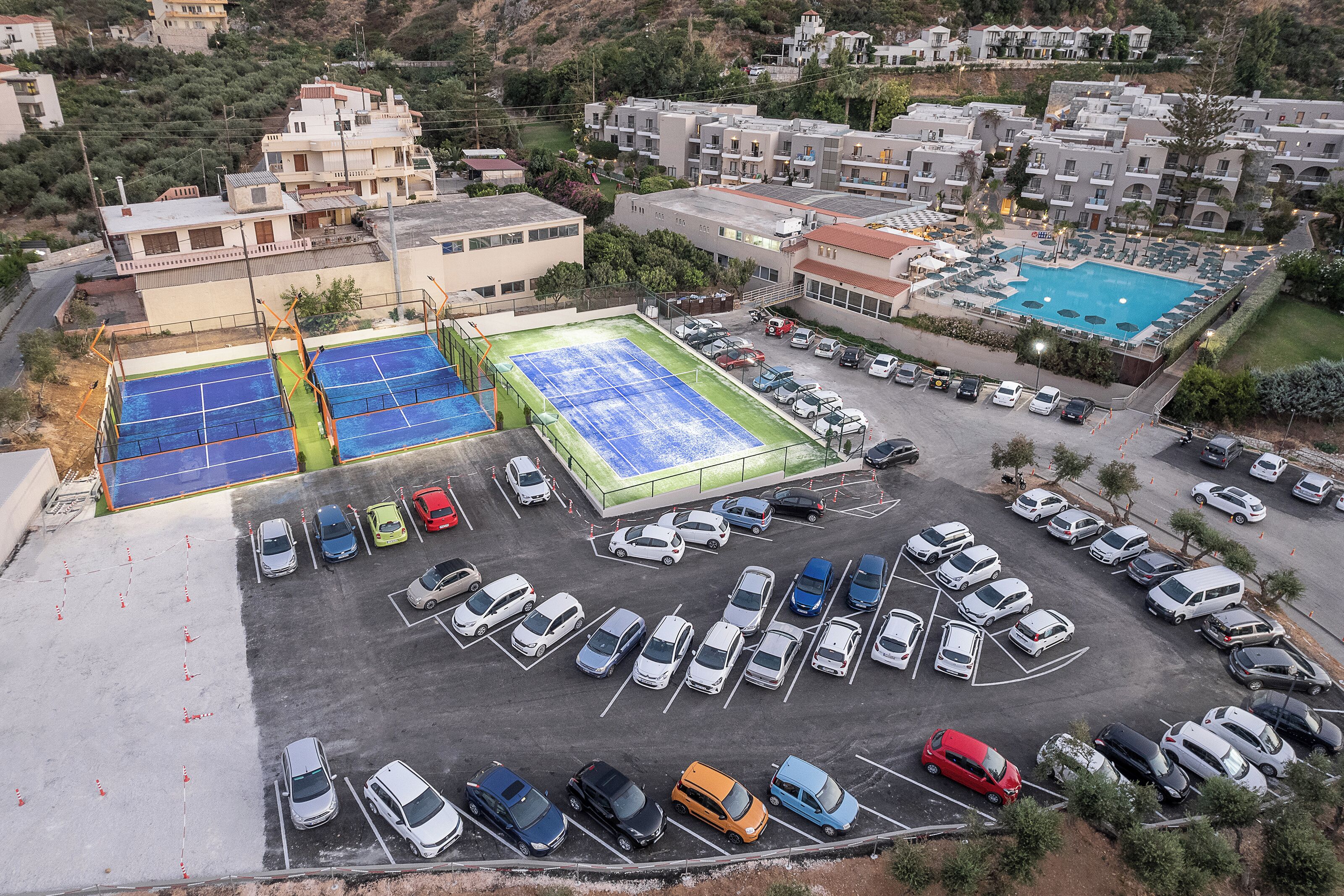 An aerial view of a parking lot with multiple cars, a tennis court, and a swimming pool.