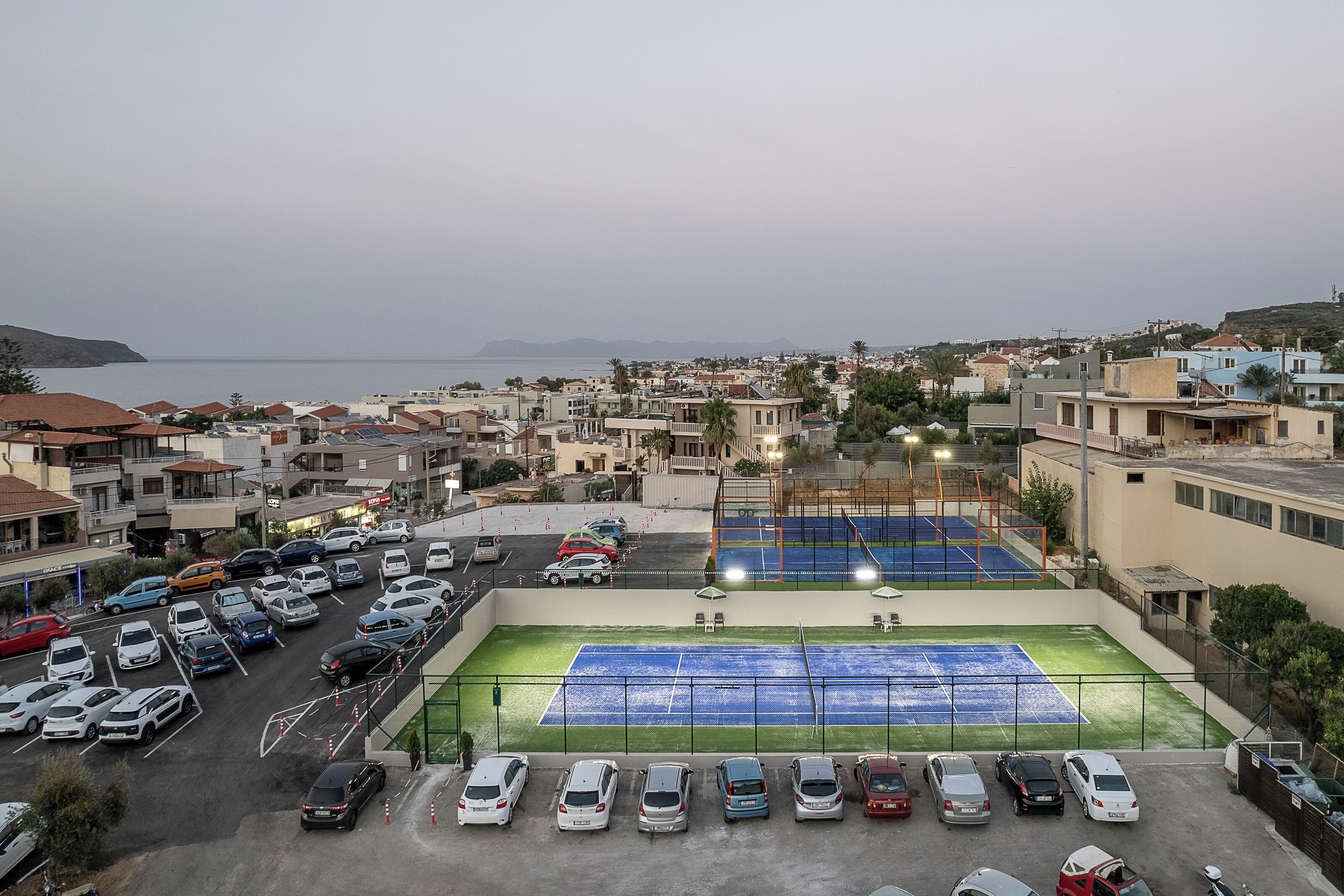 Zona residencial con cancha de tenis, varios coches aparcados y edificios con balcones.