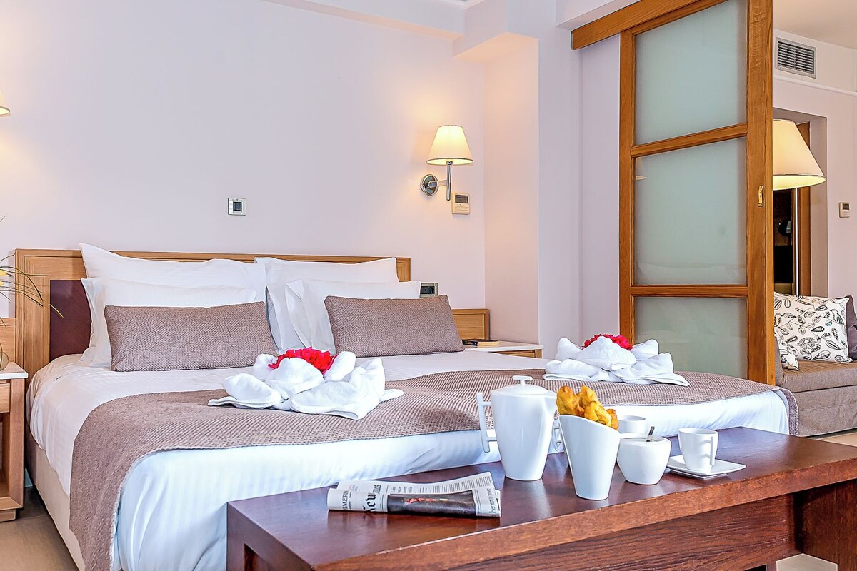 Una habitación de hotel con una cama, mesitas de noche, un pequeño mueble de madera, un banco de madera, una mesa de centro de madera y una puerta de cristal.