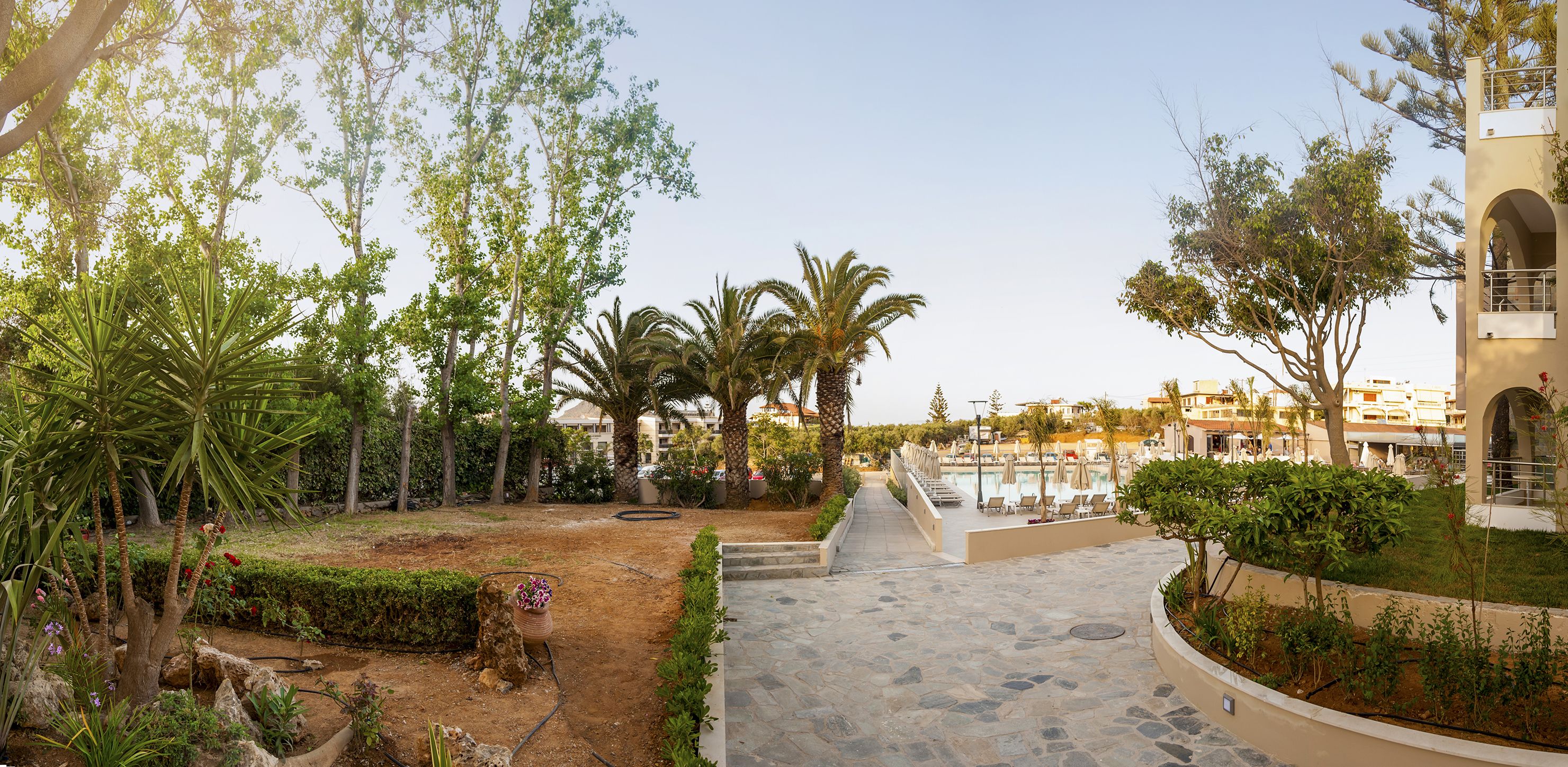 Un área exterior ajardinada con piscina, palmeras y un camino que conduce a un edificio.