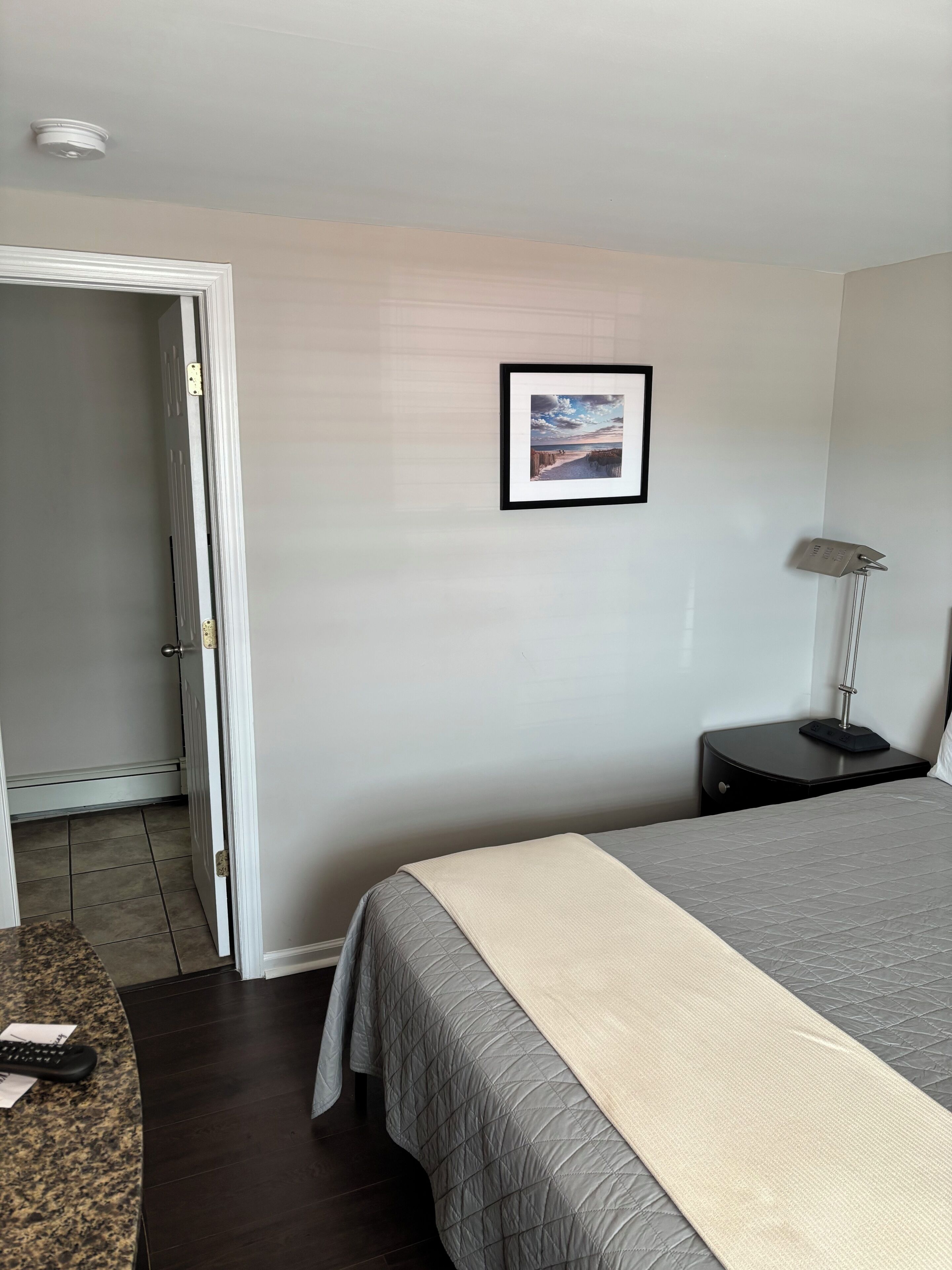 A hotel room with a bed, a nightstand, a framed picture on the wall, and a door leading to another room.