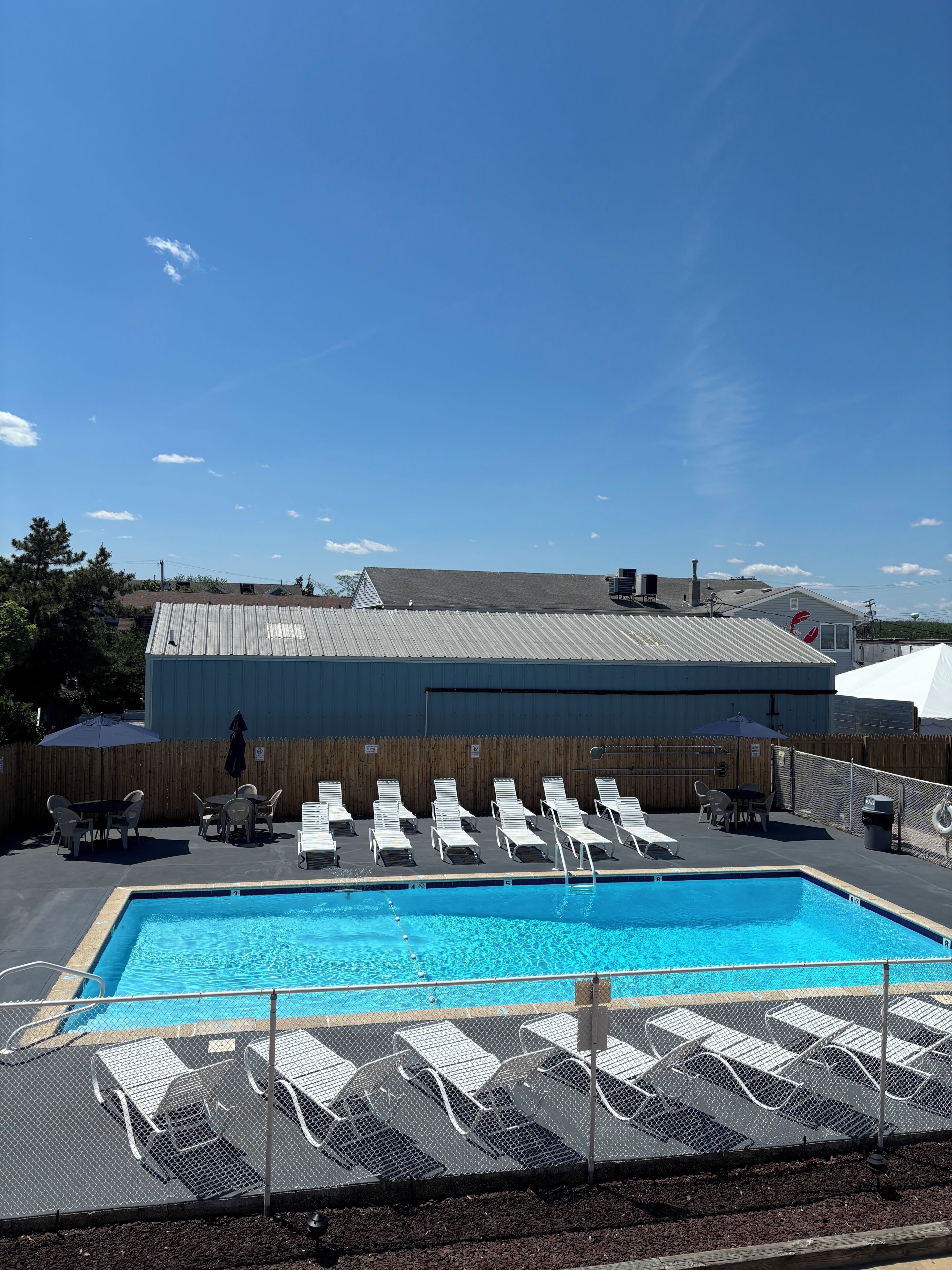 A swimming pool with lounge chairs and tables, a covered patio area, and a building with a metal roof in the background.