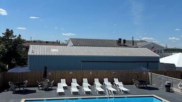A swimming pool with lounge chairs and tables, a covered patio area, and a building with a metal roof in the background.