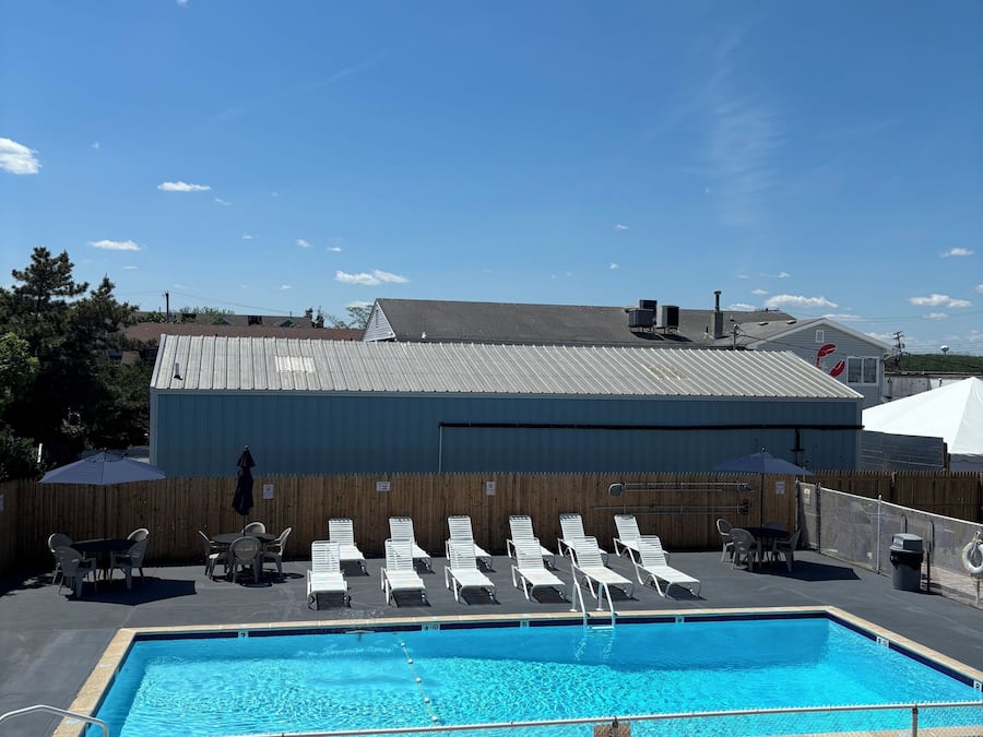 A swimming pool with lounge chairs and tables, a covered patio area, and a building with a metal roof in the background.