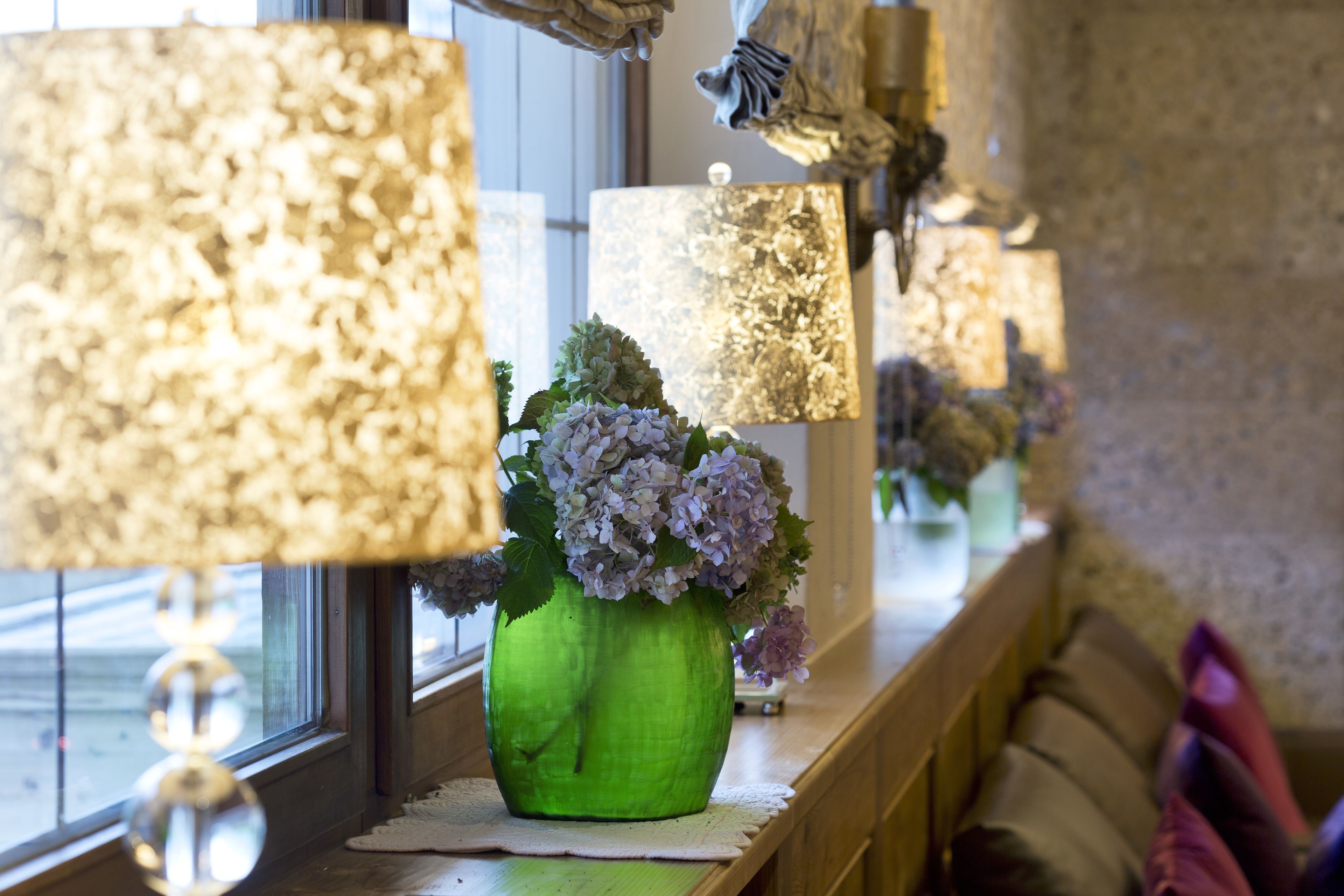 A green vase with flowers on a windowsill, surrounded by large lamps.