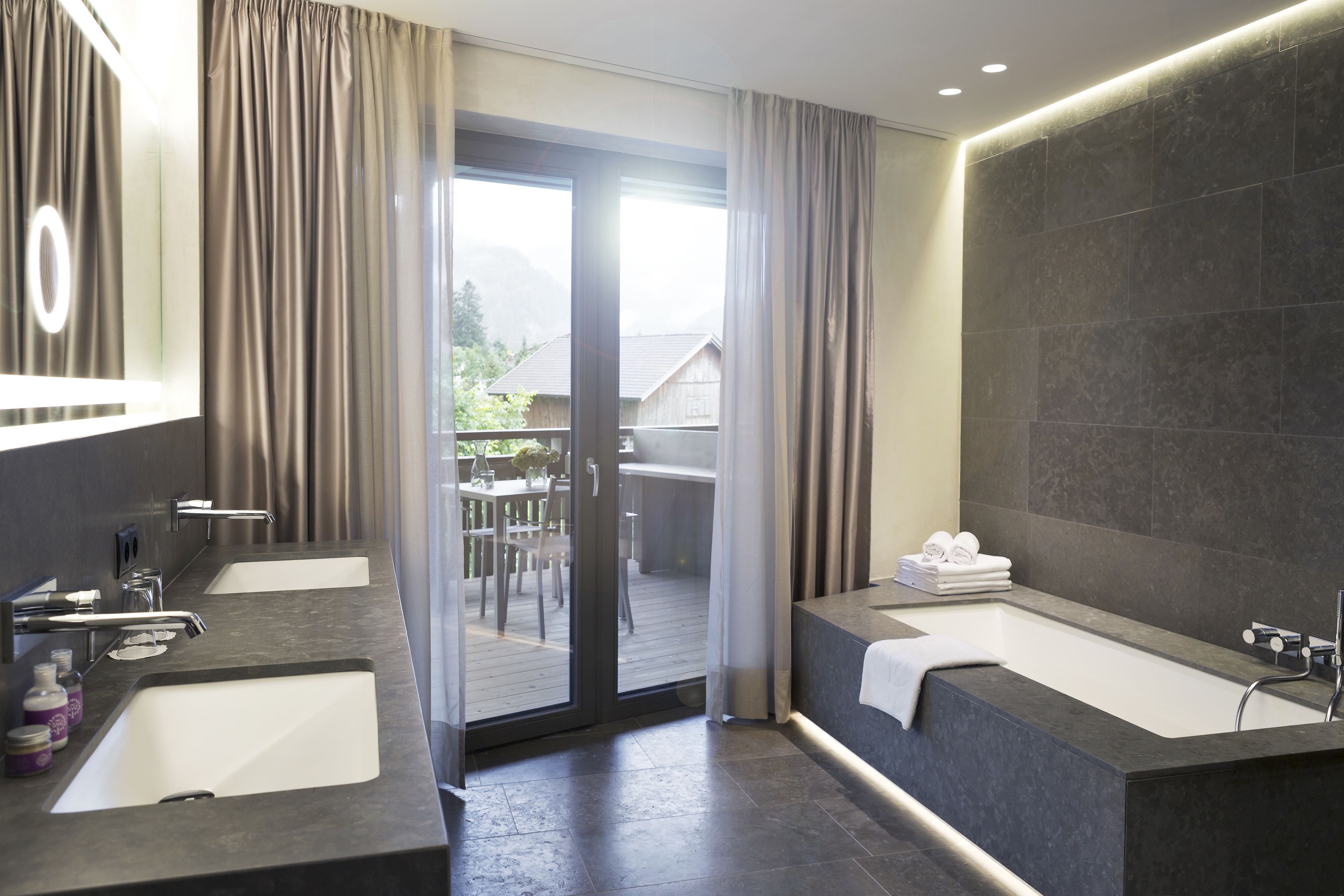 A modern bathroom with two sinks, a large bathtub, and a glass door leading to a balcony.