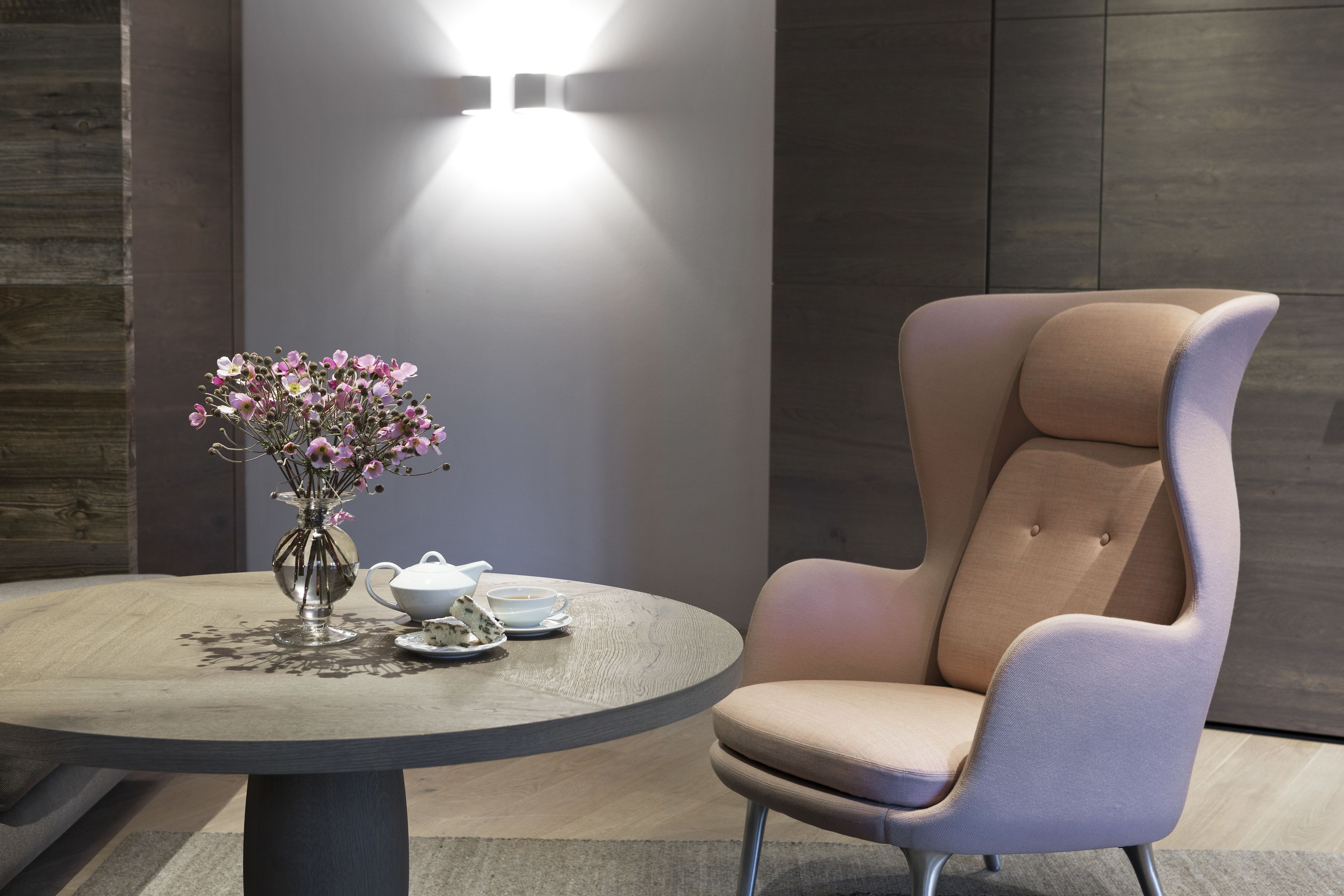 Une table ronde ornée d’un vase de fleurs, d’une théière et d’une tasse. Une chaise moderne est placée sur la droite.
