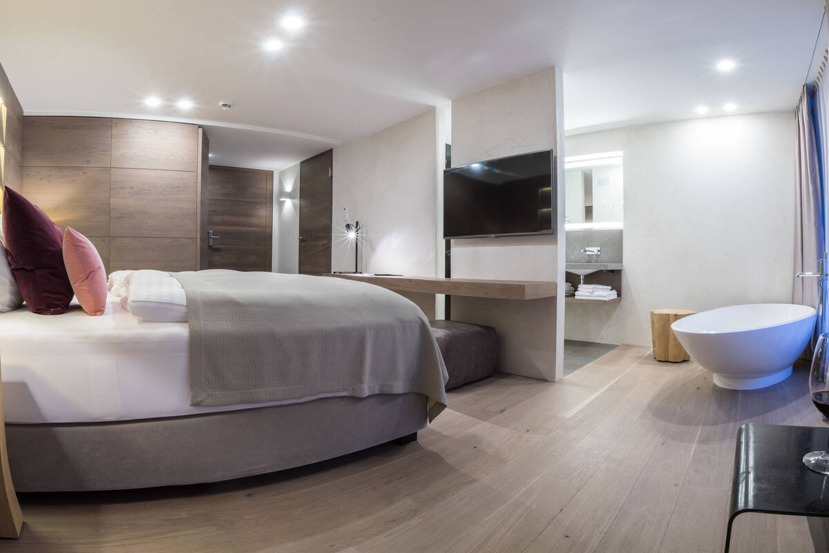 Une chambre d’hôtel moderne dotée d’un lit aux lignes courbes, d’une table de chevet, d’un téléviseur à écran plat, d’une grande fenêtre et d ’une salle de bain avec une baignoire et un lavabo.