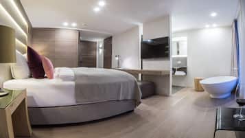 Una habitación de hotel moderna con una cama curvada, una mesita de noche, un televisor de pantalla plana, un ventanal grande y un baño con bañera y lavamanos.