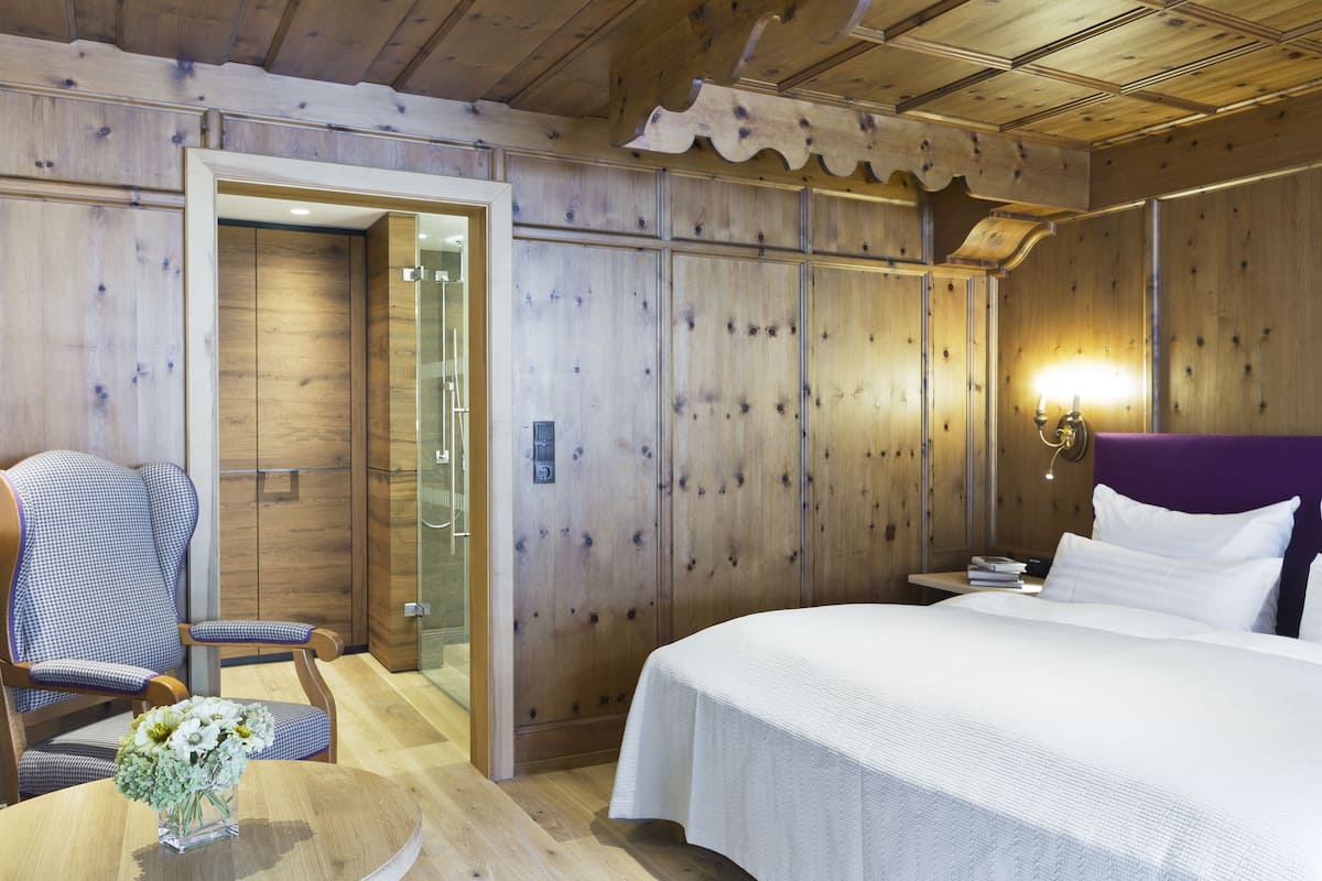 Une chambre d’hôtel avec des boiseries, un lit recouvert de draps blancs, une chaise à motifs à carreaux et une table de chevet avec des livres et une lampe.
