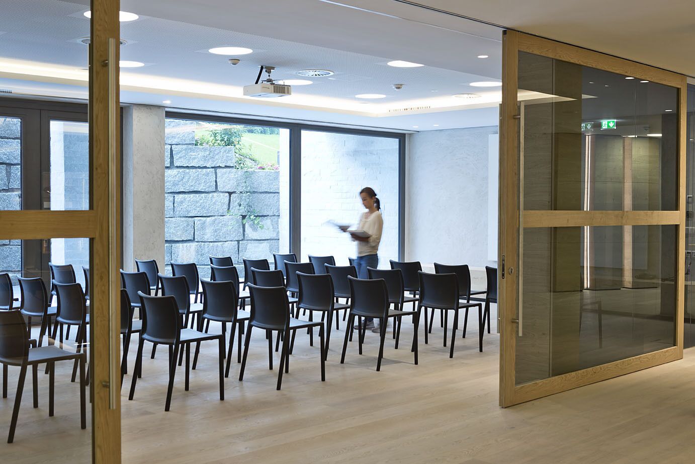 Un salón de conferencias con diseño moderno, que cuenta con un amplio ventanal, un proyector y una persona de pie cerca de la ventana.