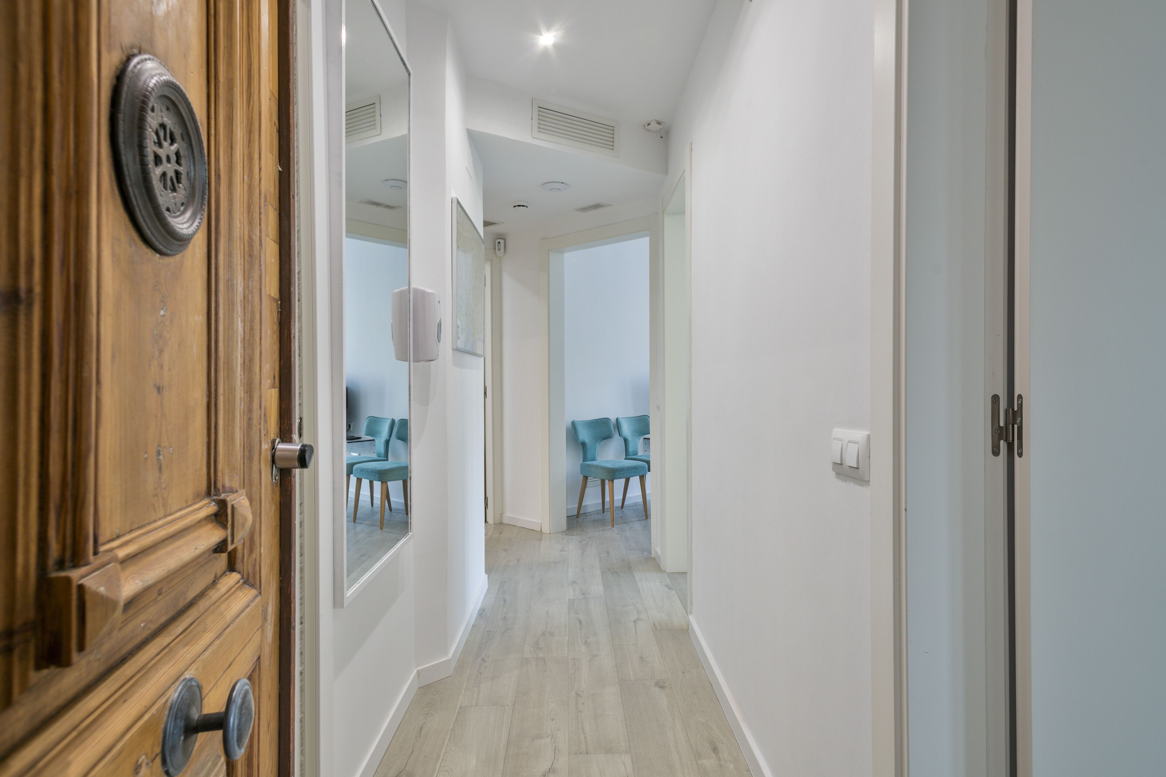A hallway with a wooden door, a light switch, and a mirror reflecting a room with two chairs.