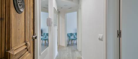 A hallway with a wooden door, a light switch, and a mirror reflecting a room with two chairs.