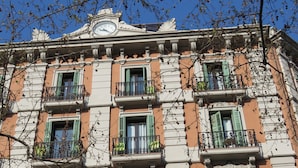 Um edifício de vários andares com detalhes arquitetônicos elaborados, incluindo um relógio, varandas com grades de metal e janelas com persianas.