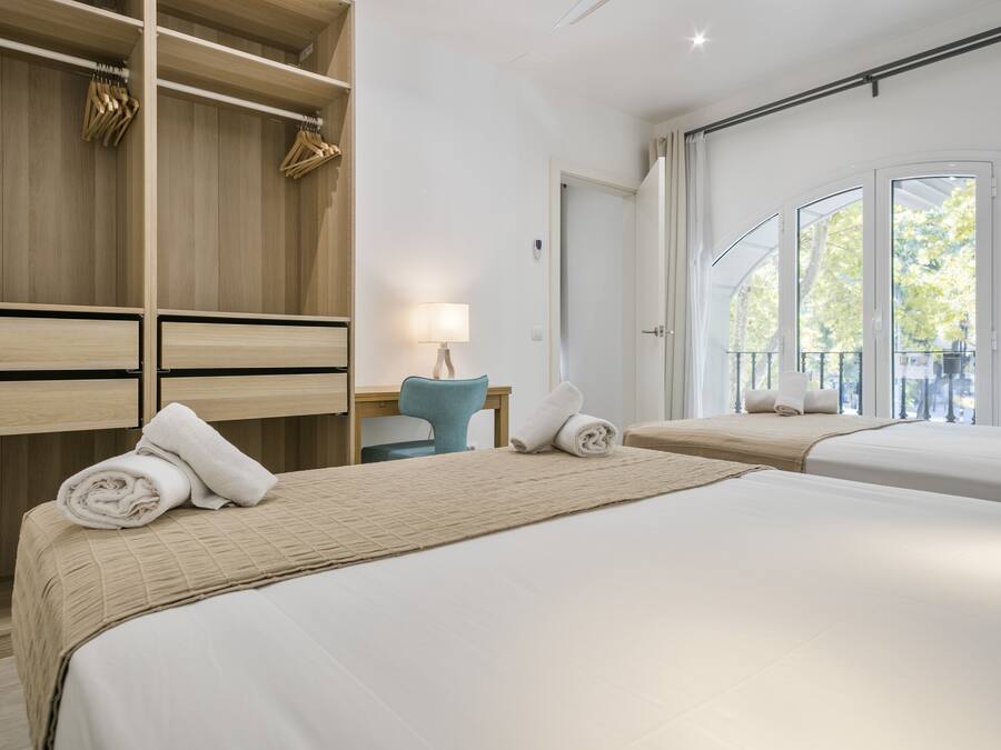 Habitación de hotel con dos camas, un armario de madera, un escritorio con lámpara y un balcón con vista a árboles.
