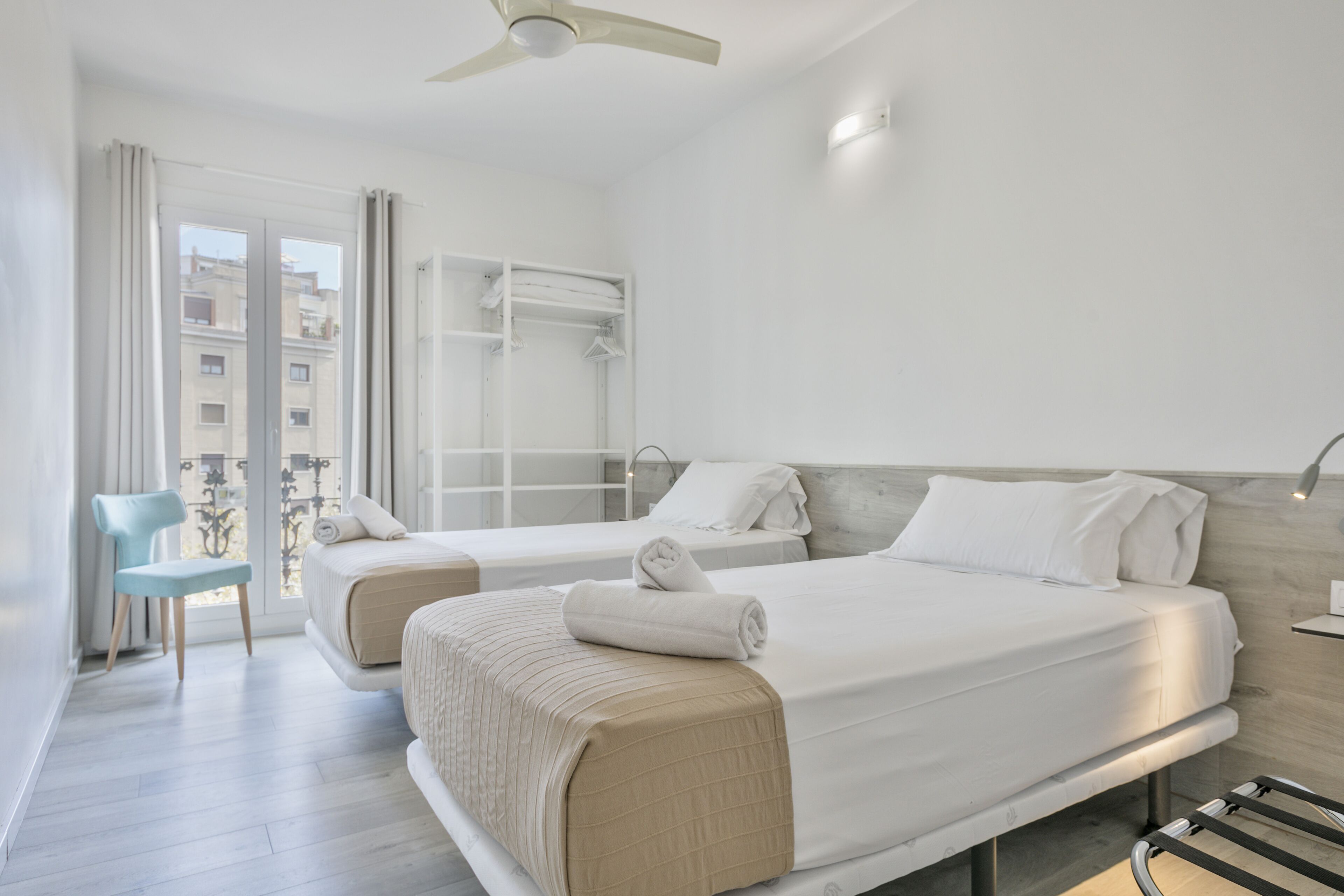A hotel room with two beds, a chair, a wardrobe, and a balcony with a view of buildings.