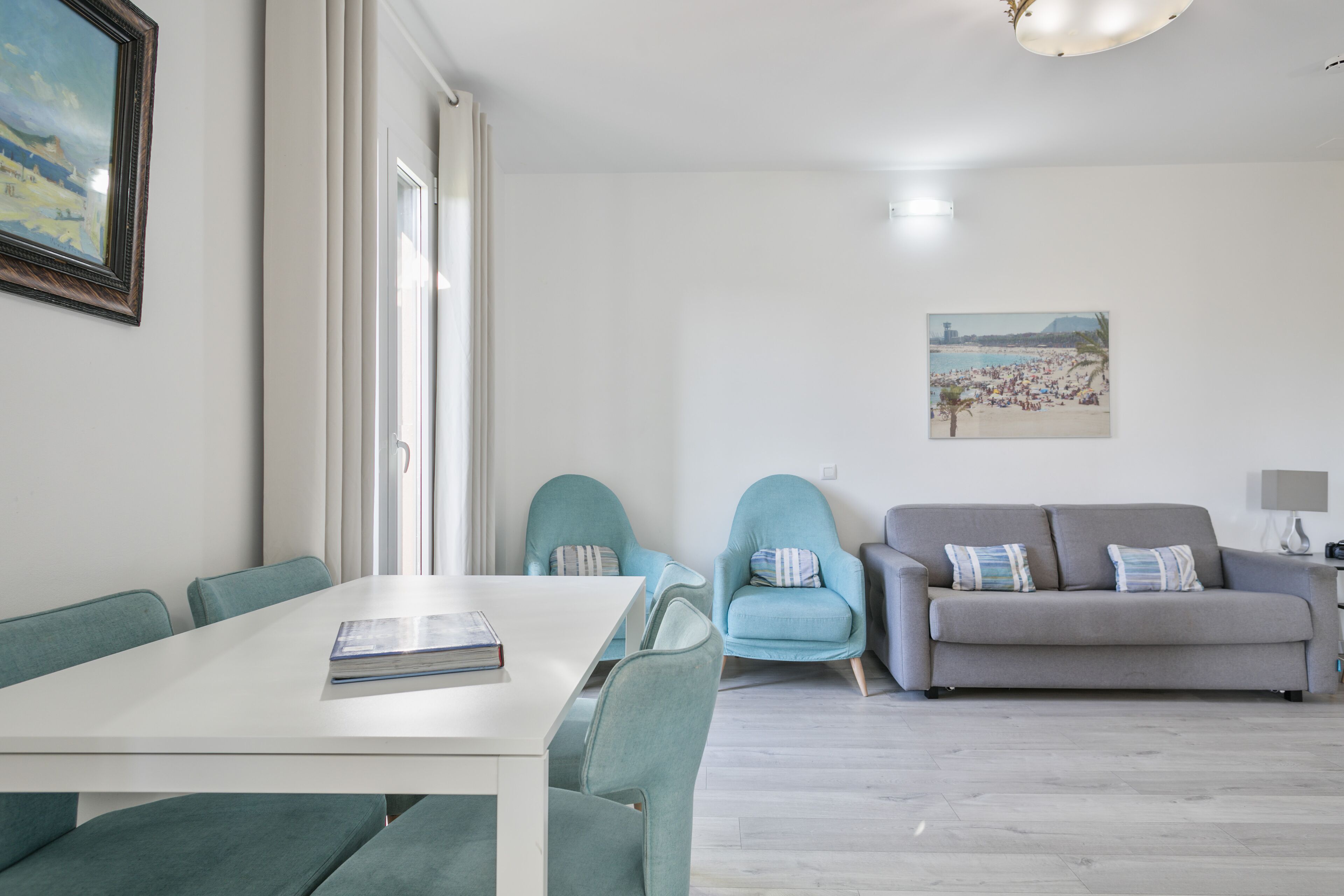 A modern living room with a white table, teal chairs, a grey sofa, and a beach-themed painting on the wall.