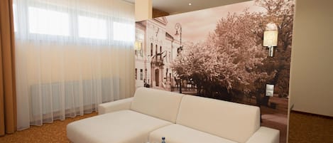 A hotel room with a large sofa, a small table with a bottle and glasses, a large window with sheer curtains, and a wall mural of a tree and buildings.
