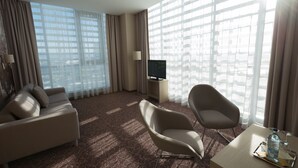 A hotel room with a large window, a sofa, a chair, a TV, and a small table with a bottle and glass. - Košice Hotel (Kosice)