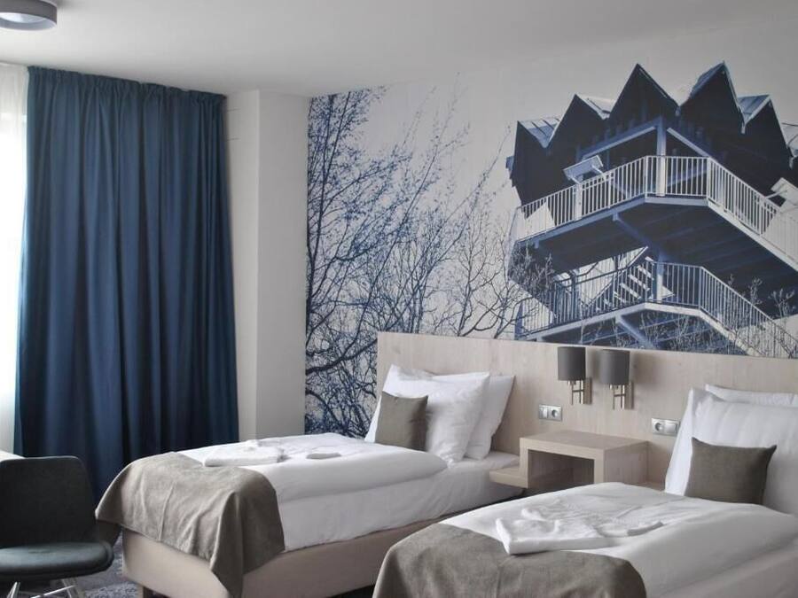 Habitación de hotel con dos camas, un escritorio y una silla. Se observa un ventanal grande con cortinas azules.
