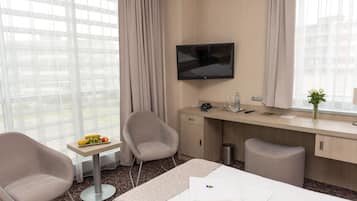 A hotel room with a bed, two armchairs, a desk with a TV, a small table with fruit, and a window with curtains.